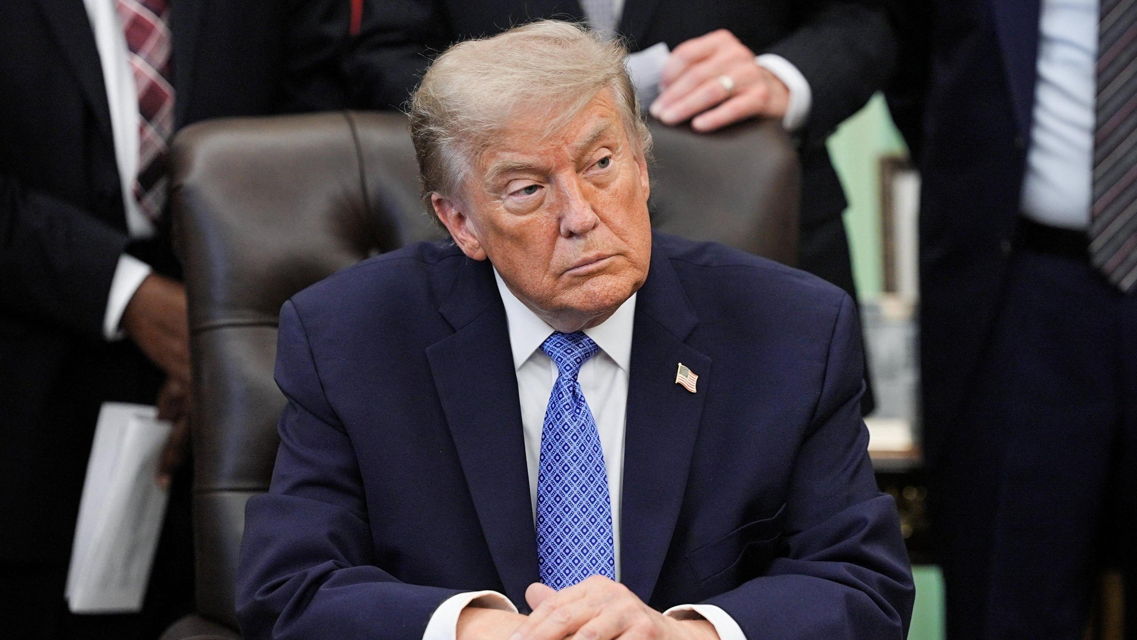 U.S. President Donald Trump sits at his desk in the Oval Office turning to his left. He is wearing a blue suit, white shirt and blue tie. He is not smiling. 