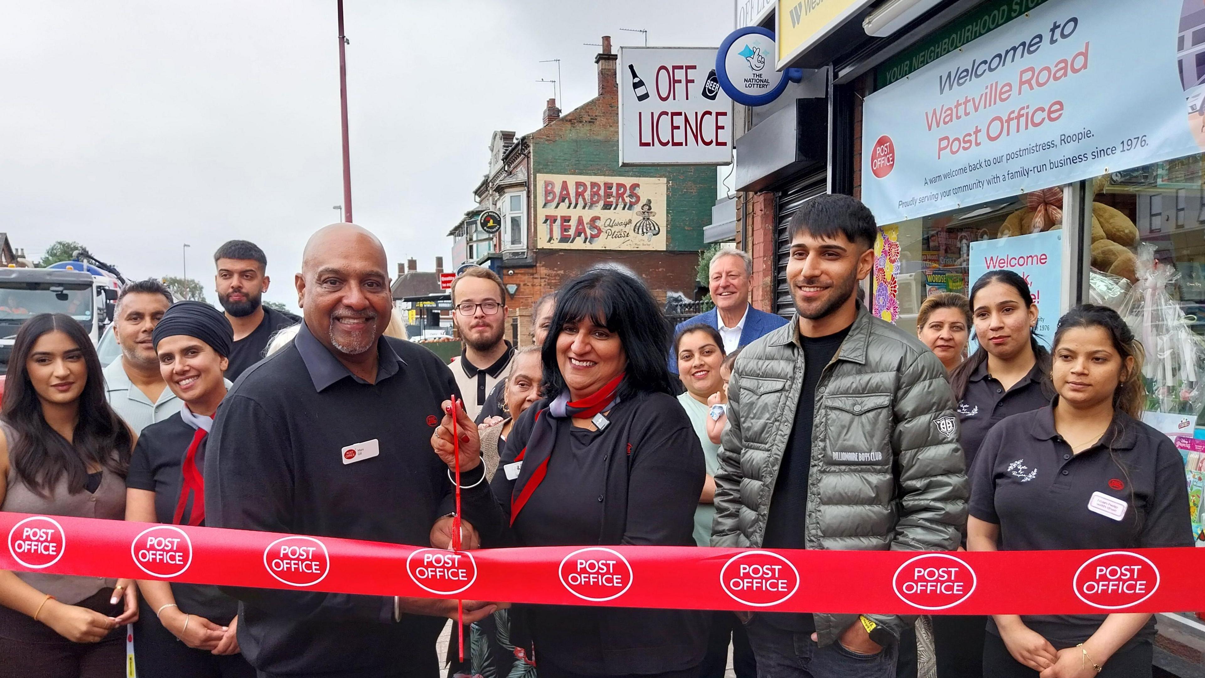 Rooprit Gill dressed in black clothing and a red and black scarf is seen cutting a red ribbon with the Post Office logo on with red scissors. She is stood with a crowd of other people outside the Post Office. 