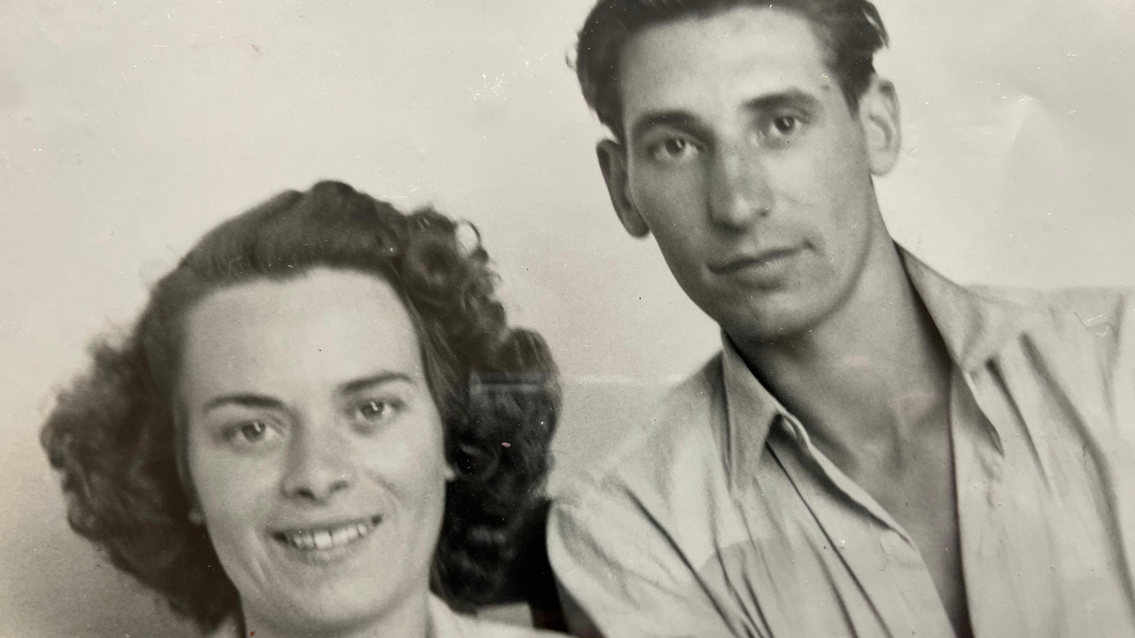 Black and white picture of a couple with a white background the lady on the left has dark black hair and a light coloured top on the man on the right also has dark hair swept across his head and an open topped light shirt.