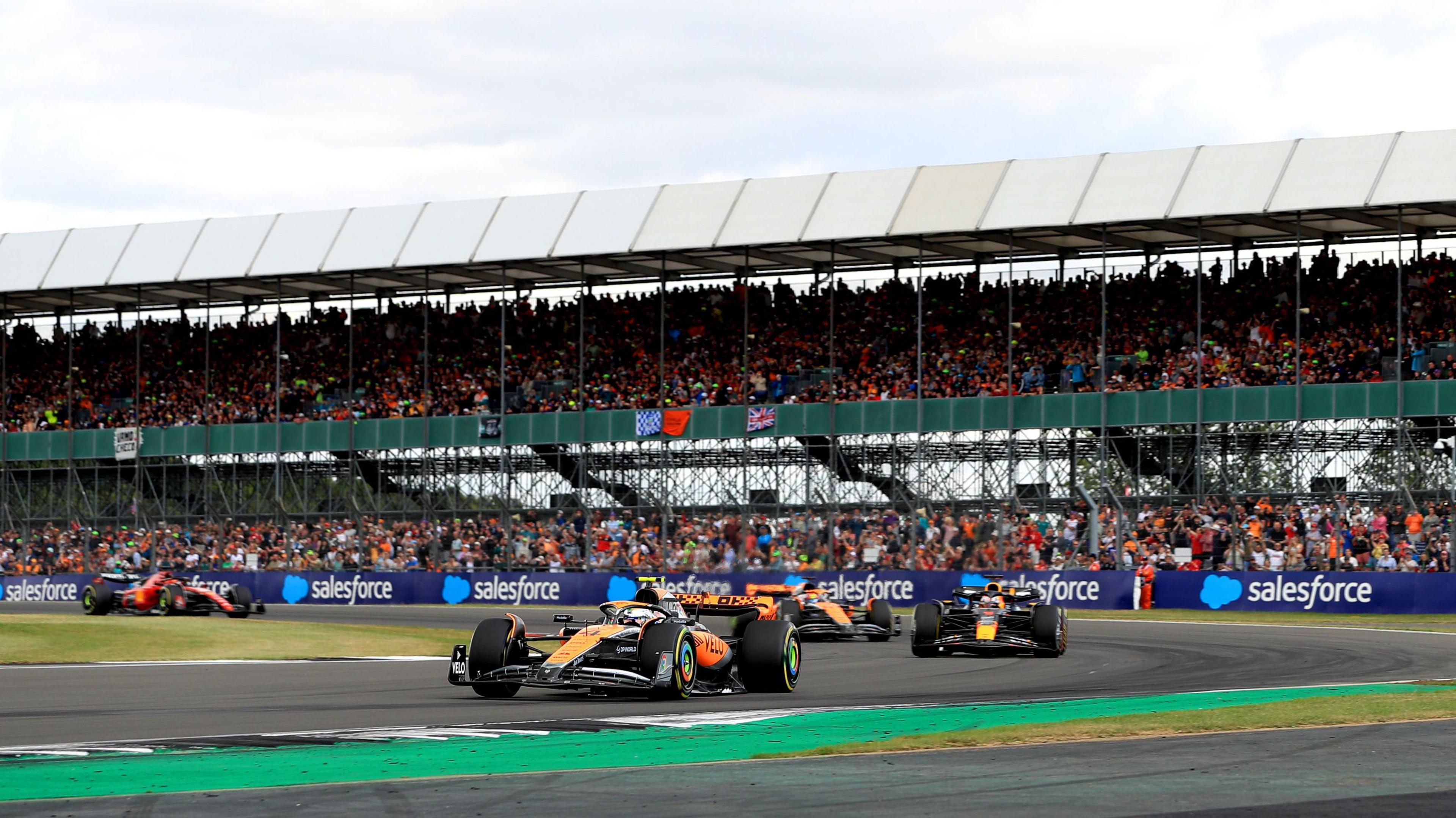 Formula 1 cars racing during the British Grand Prix at Silverstone. Cars from McLaren, Red Bull and Ferrari are all shown against the backdrop of a packed grandstand.