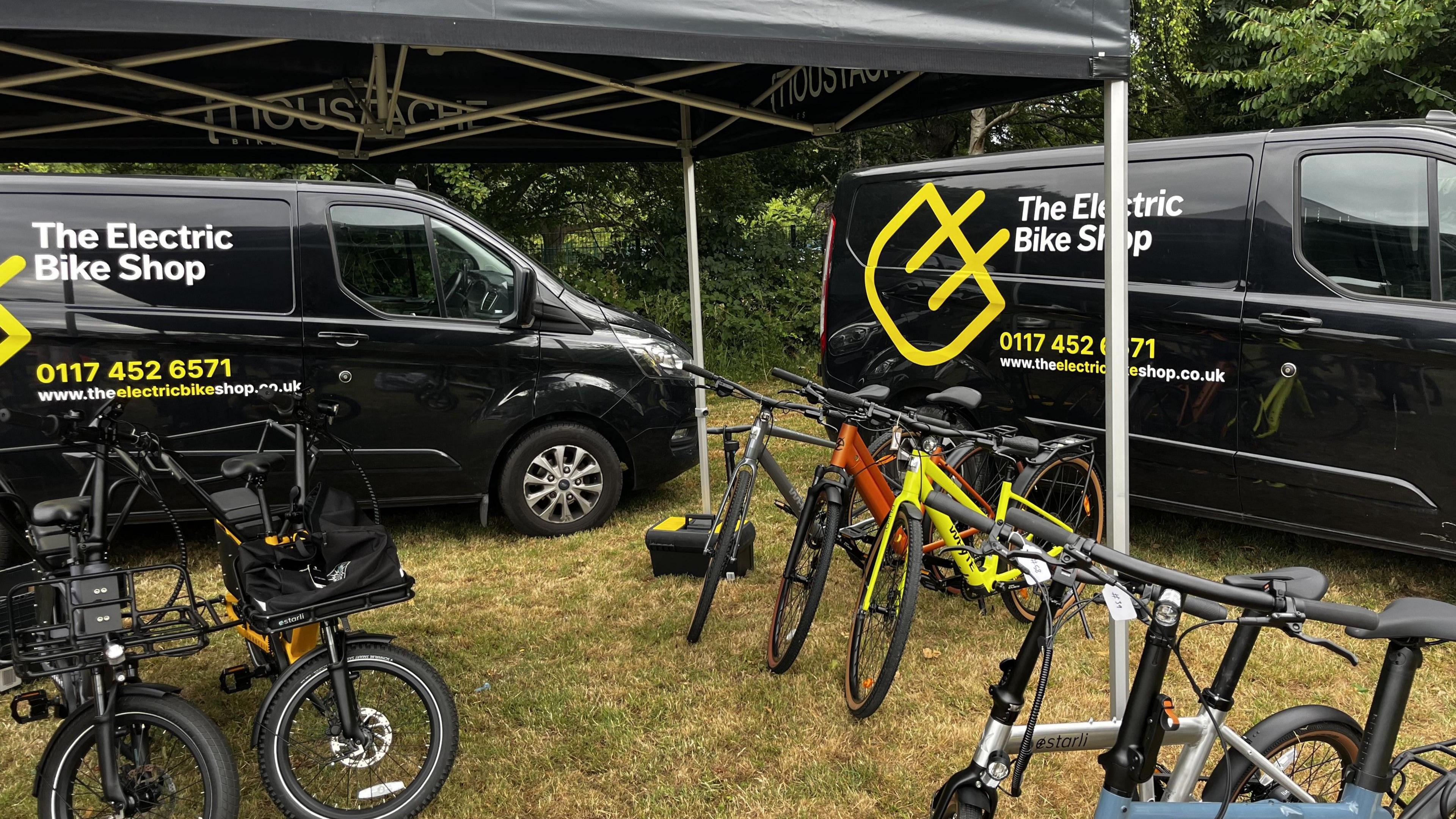 The image shows two black vans at the back of the image with 'The electric bike shop' logo in yellow. In front of the vans are seven different types of electric bikes.