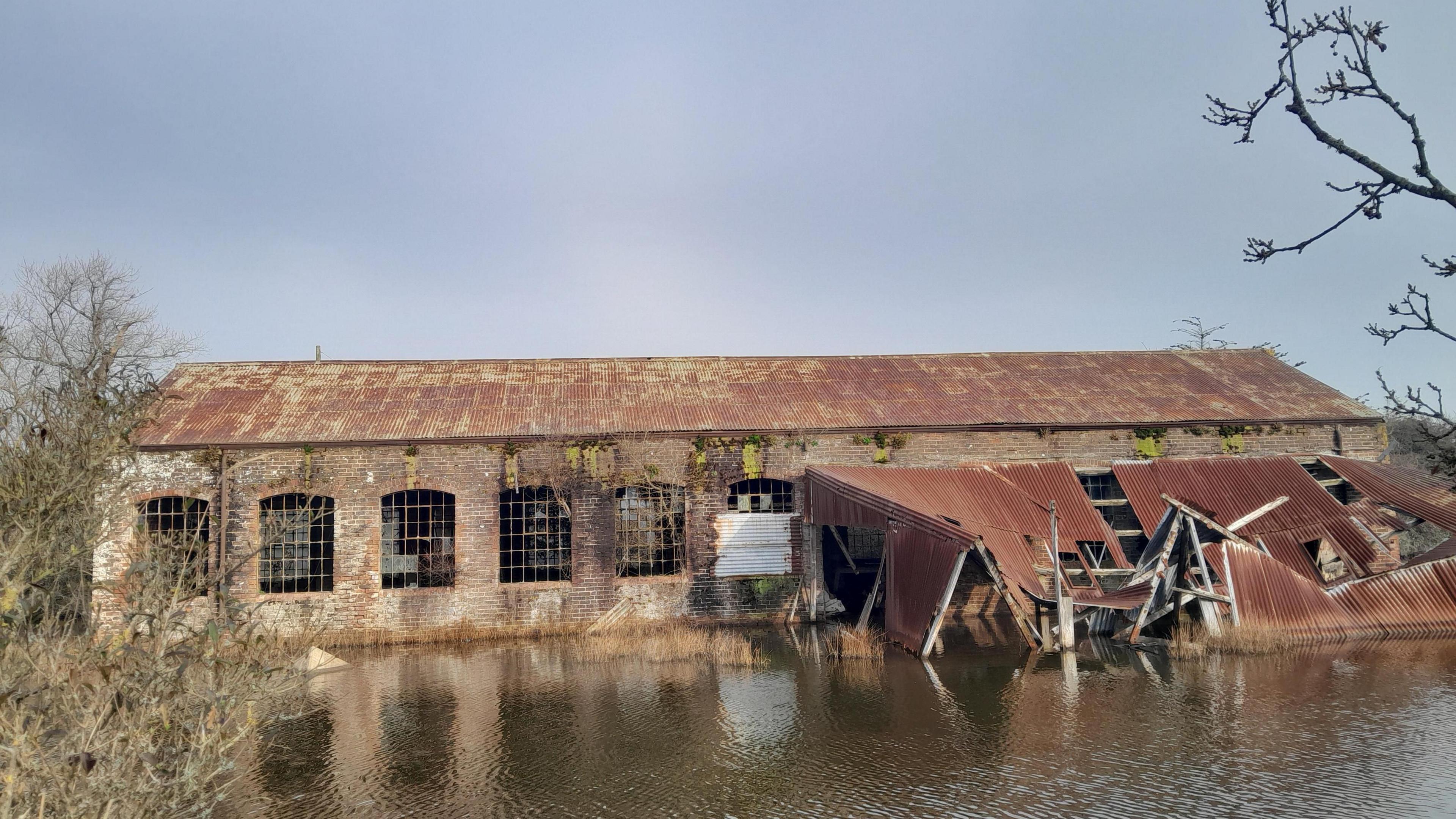 A large derelict warehouse building with windows and a corrugated roof stands by a body of water.  The building appears to built of bricks. Part of the structure has collapsed into the water. The sky overhear is grey.