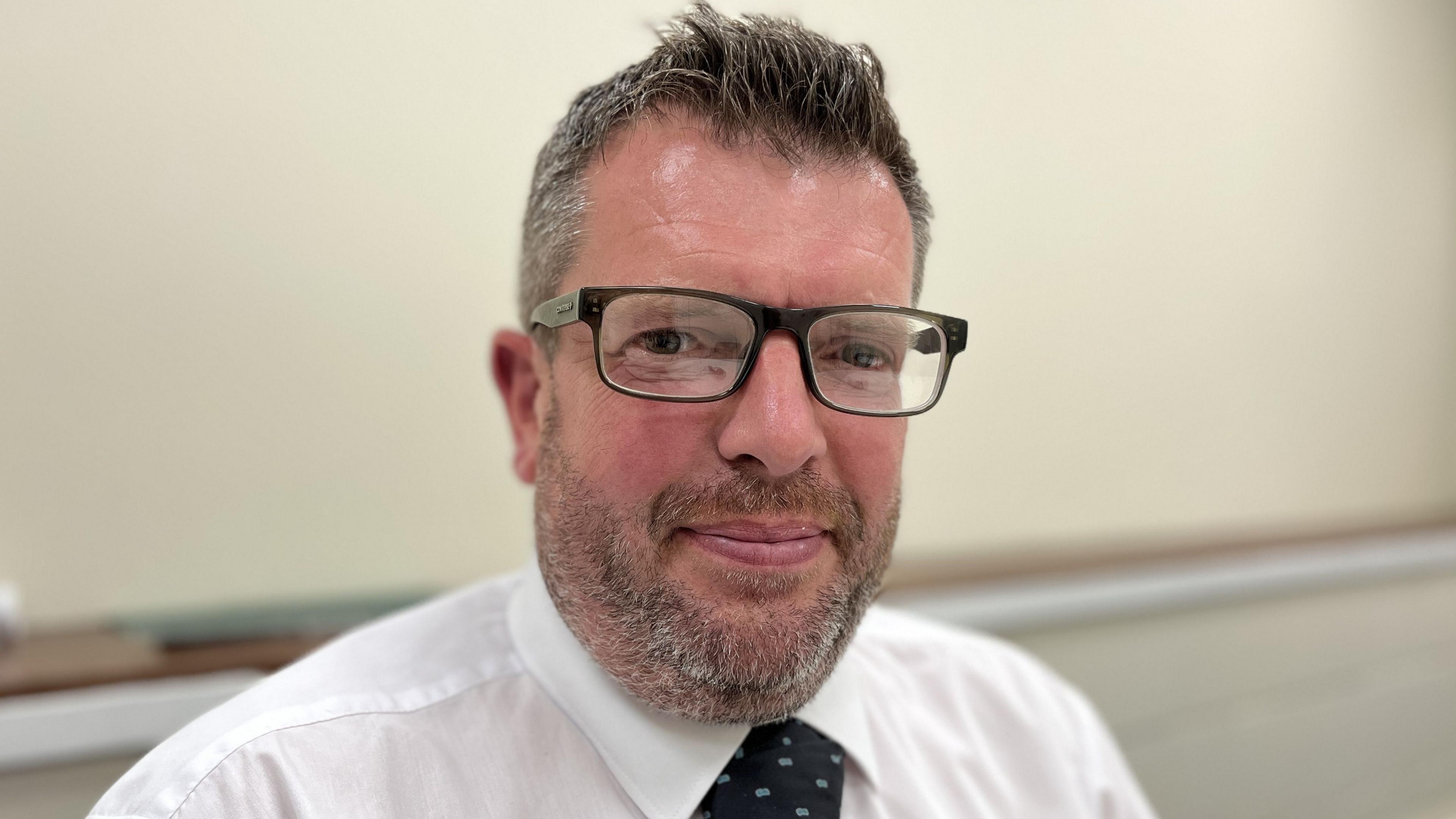 Luke Barnes with black rimmed glasses short grey hair and beard wearing a white shirt and tie