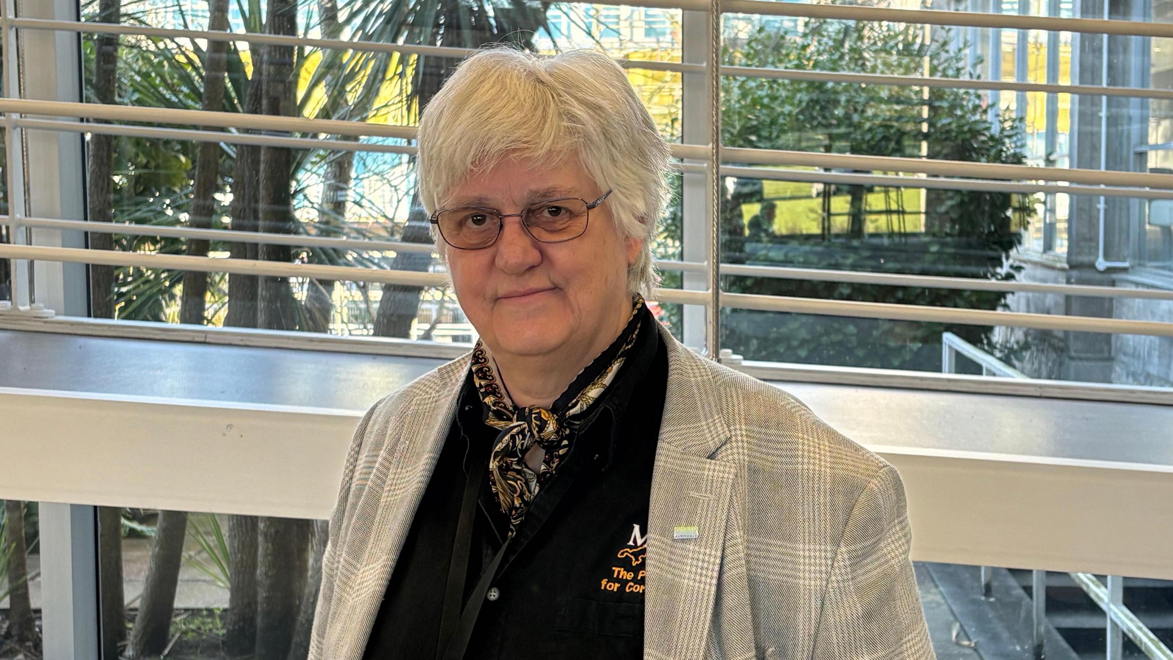 Loveday has short white hair and glasses and wears a light grey checked jacked and black top. She stands in front of a walkway and windows at County Hall. 