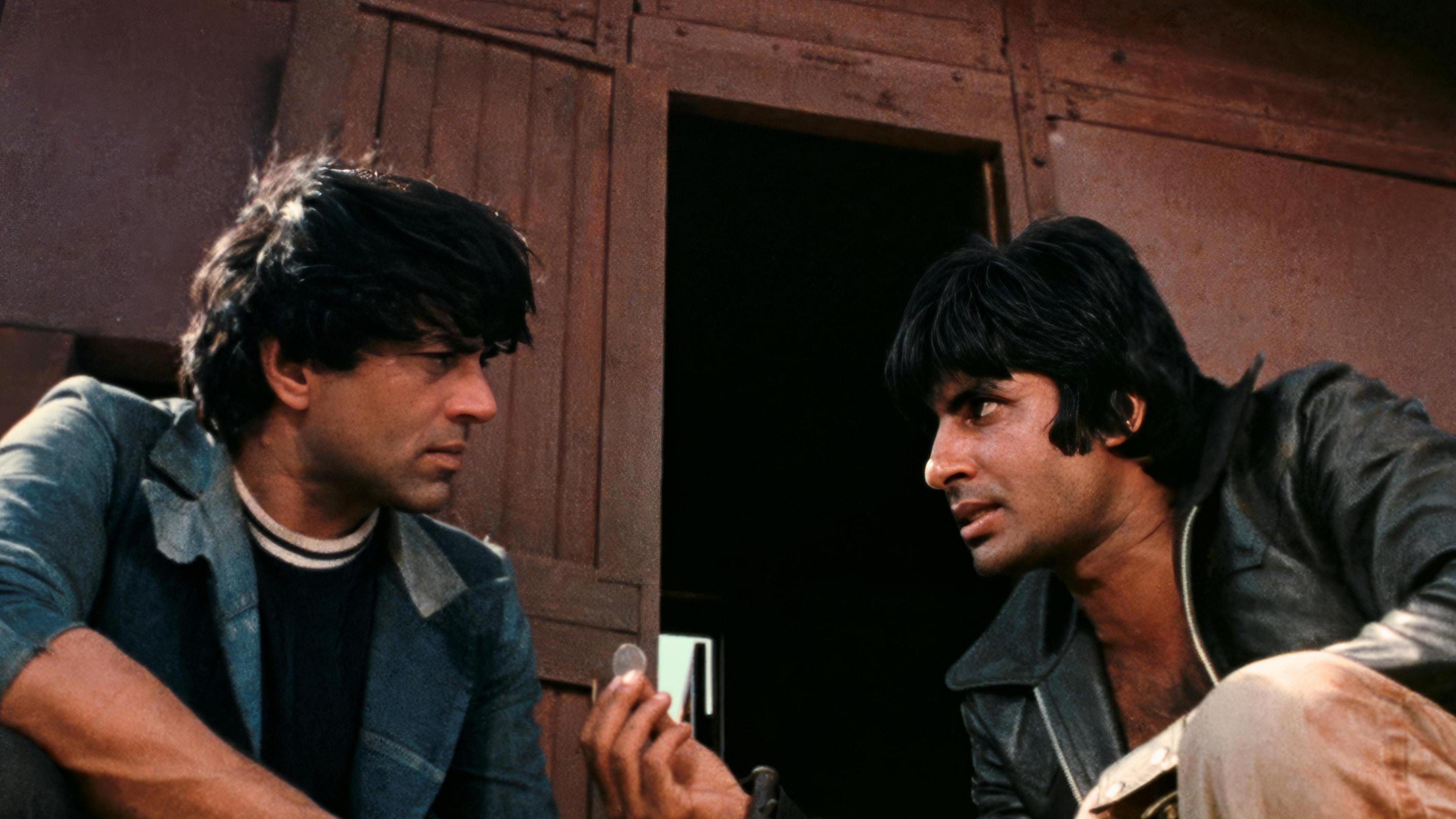 The image shows the two leading men of Sholay Amitabh Bachchan (right) and Dharmendra (left) wearing jackets and engaged in a conversation.