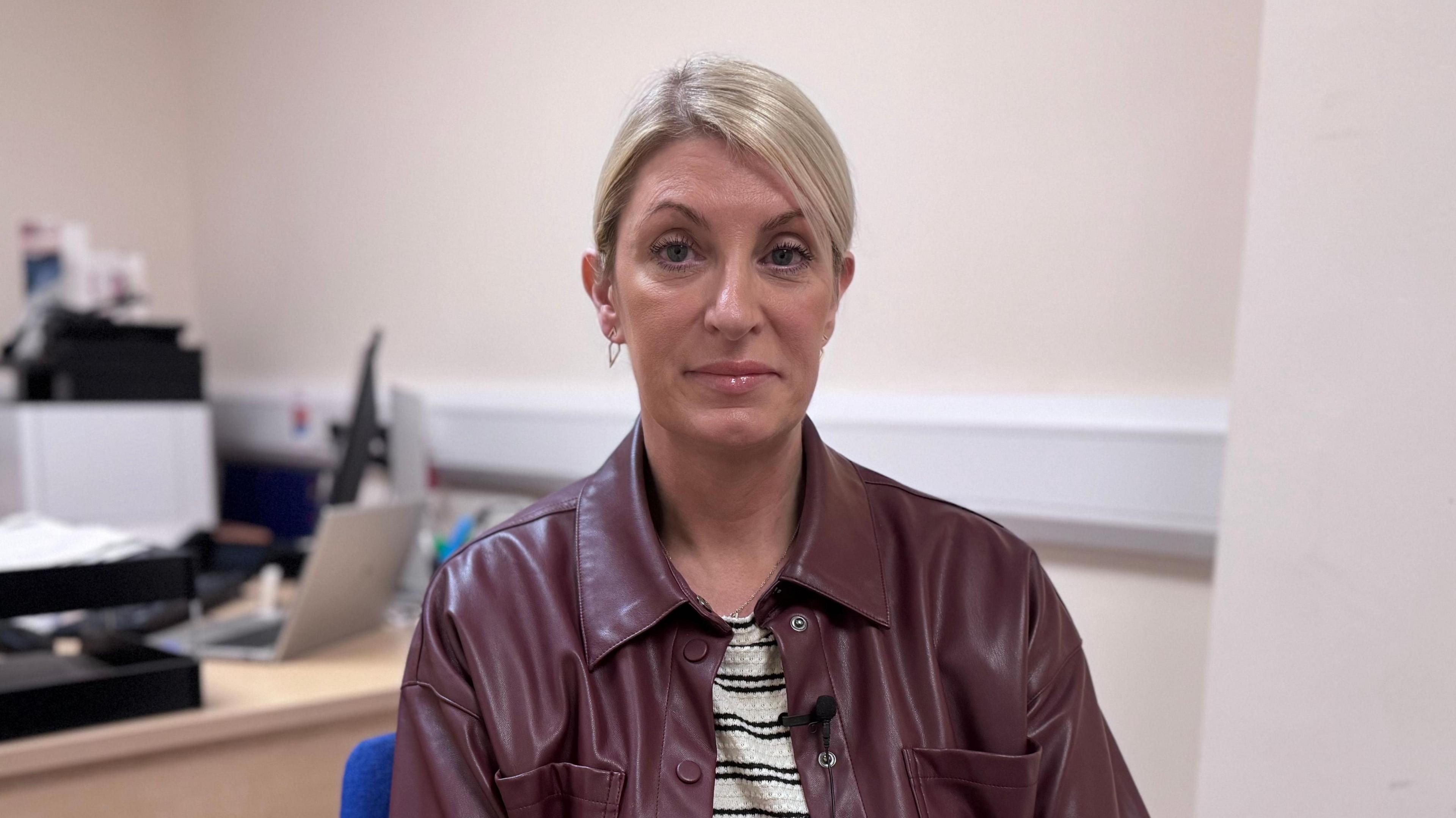 Linda Dillon. She has blonde hair tied back, wearing a brown leather shacket and black and white striped top.