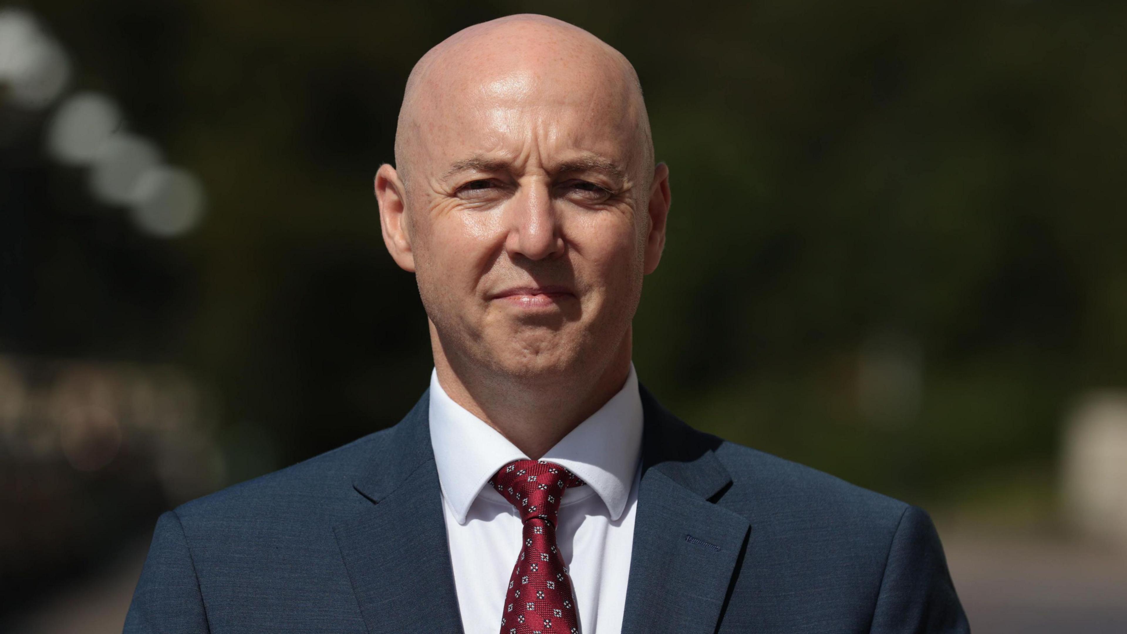 Jon Burrows looking at the camera. He is wearing a suit that is charcoal with a white shirt and a red tie.