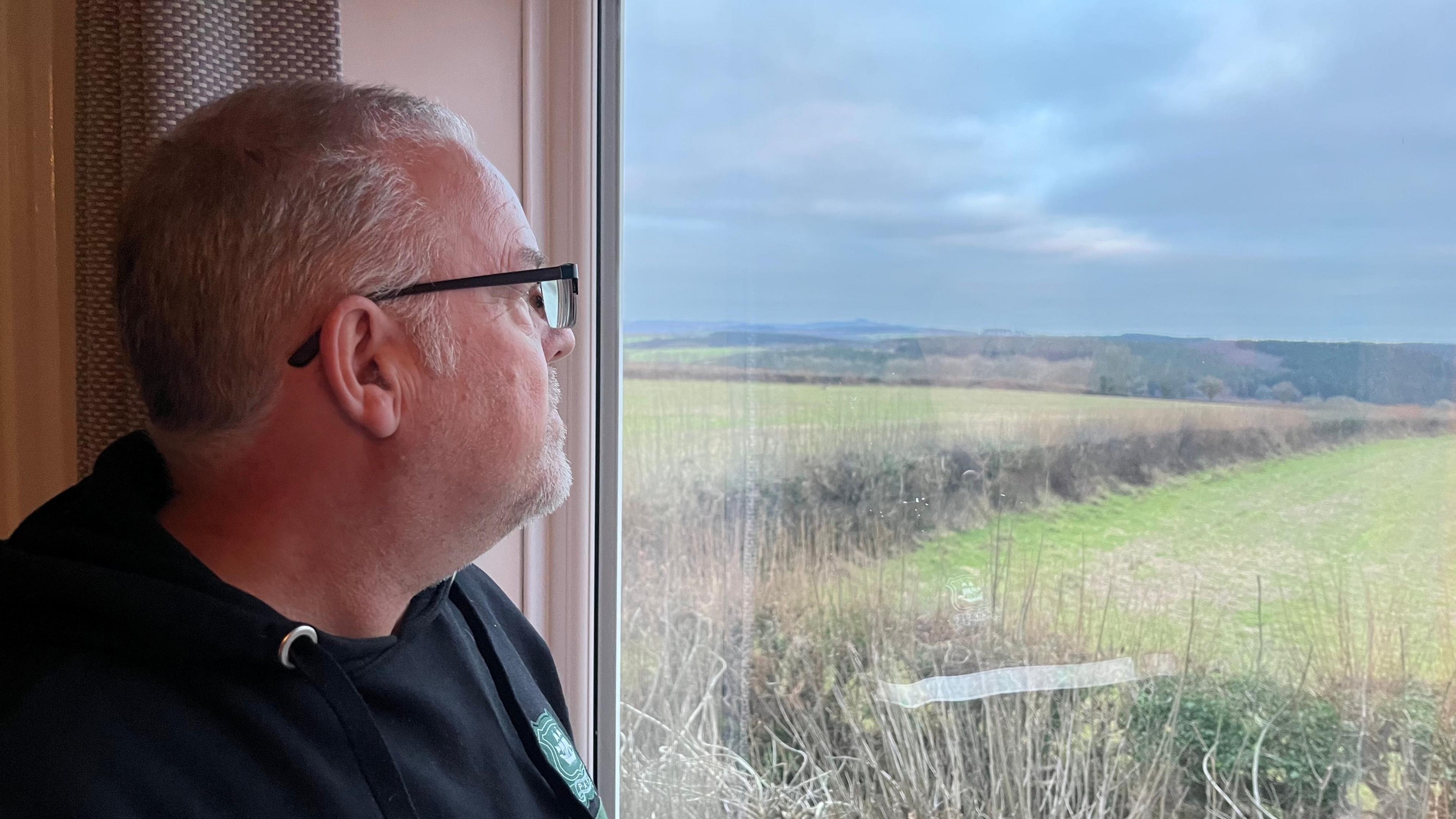 Steve Newell, Woolwell resident, wearing a black hoodie and looking out of his window over fields towards Dartmoor