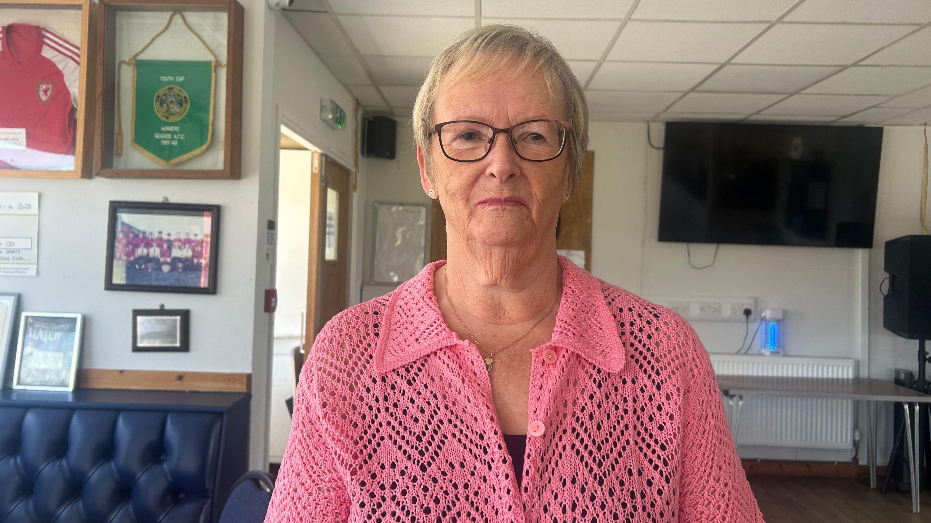 Julianna Millard, 70, mewn cardigan pinc a'n gwisgo sbectol. Mae ganddi wallt byr melyn a mwlcus aur. 