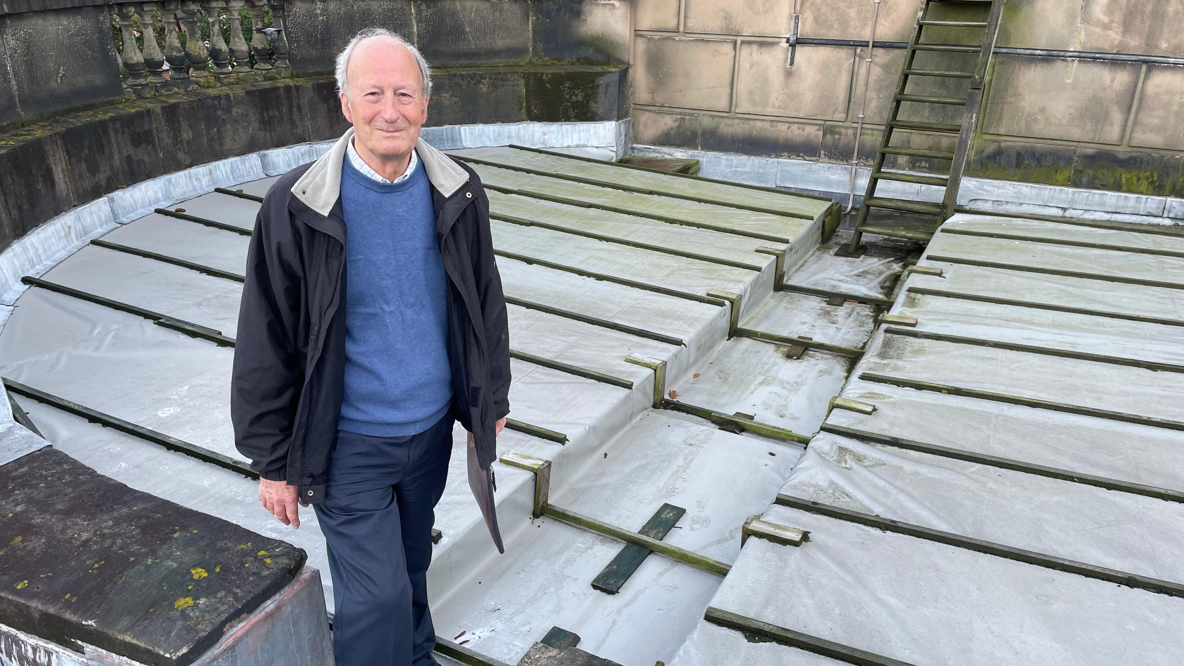 Ian Somervaille is standing on a church roof. There is white plastic covering over the section of roof.