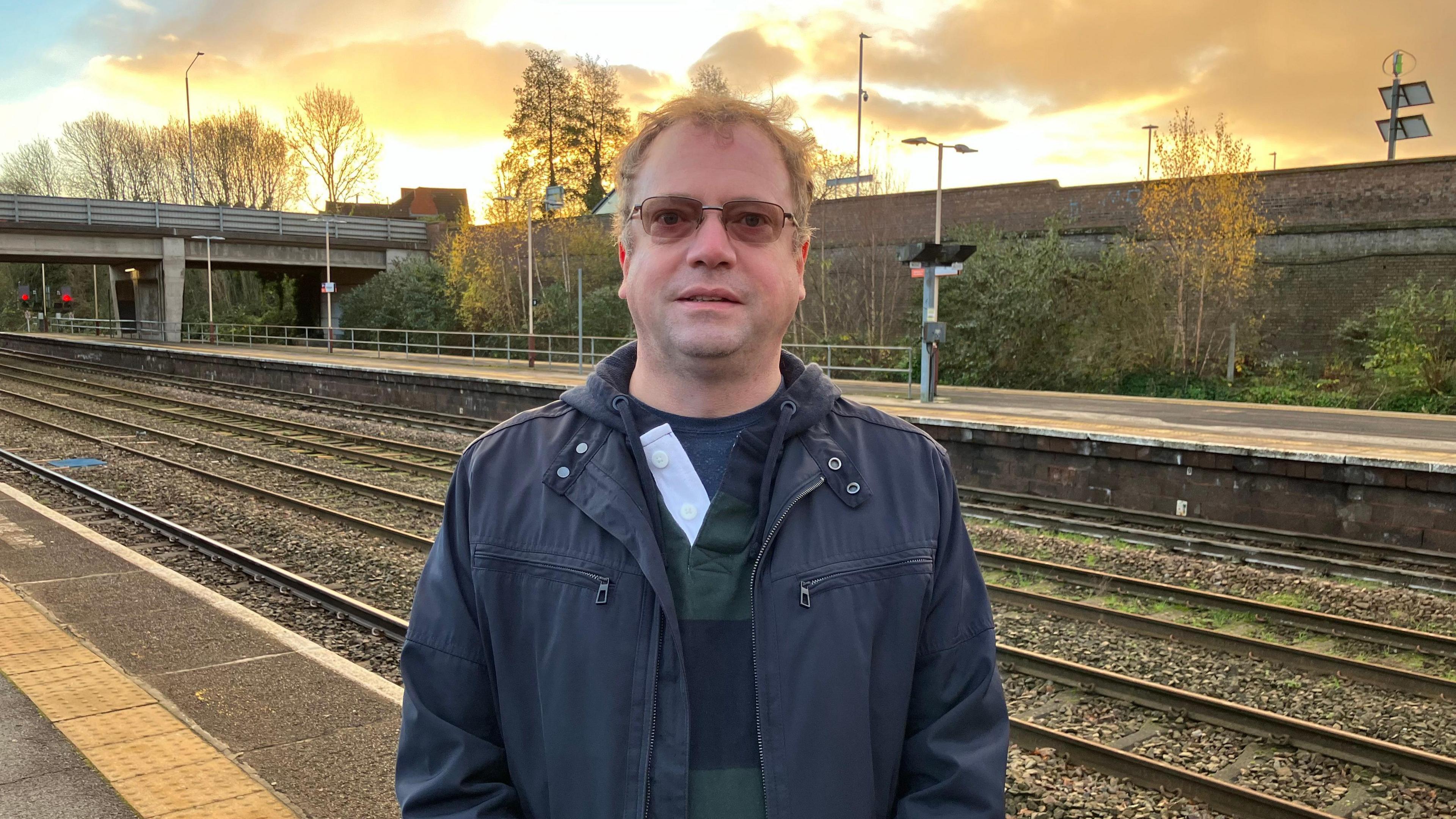 Chris Taft is standing on a train platform with railway lines in the background. He has light brown hair and wears tinted glasses. He has a navy blue jacket on, with a navy and dark green striped top. The sky in the background is golden yellow, which illuminates the clouds.