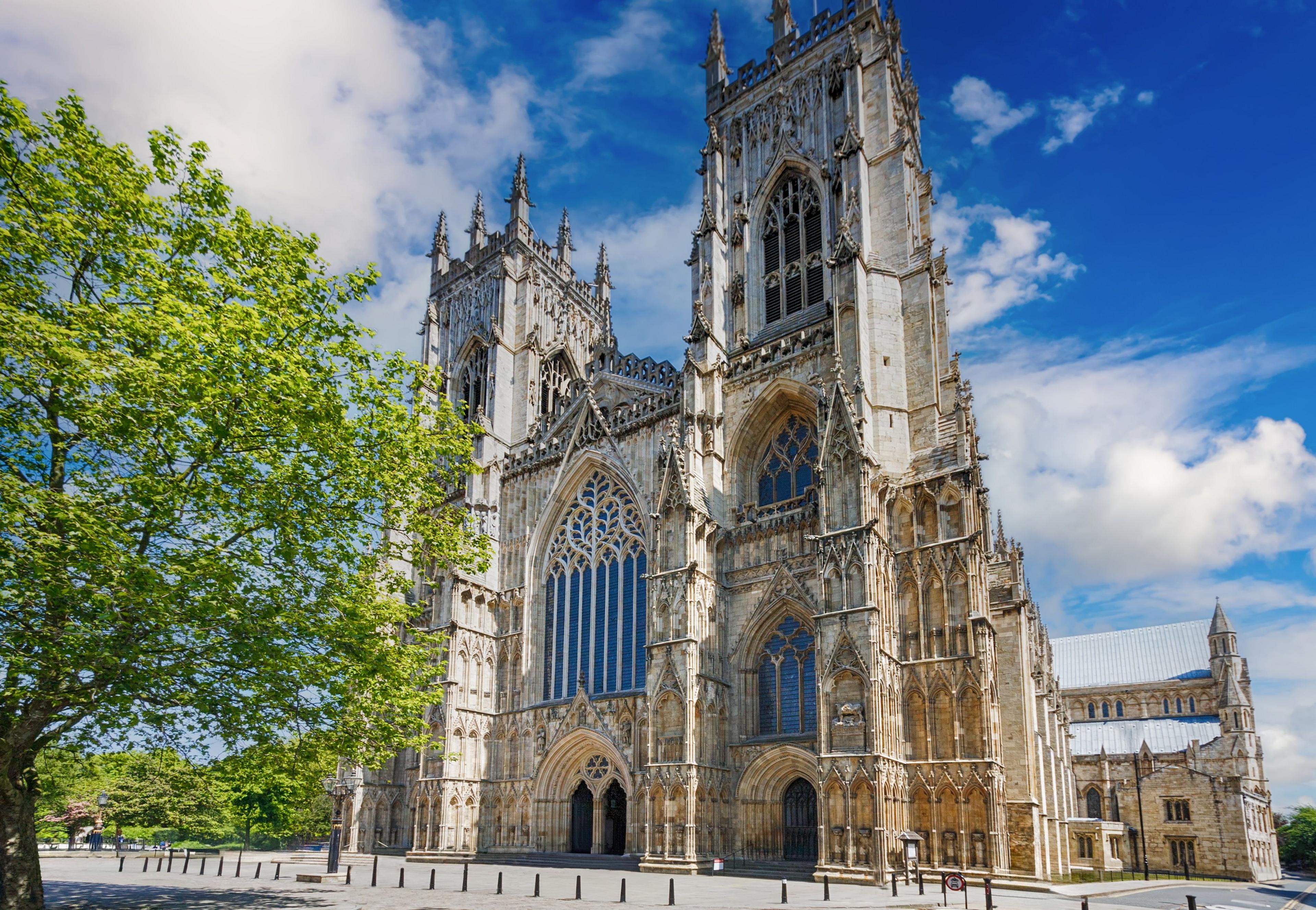 York Minster is a large gothic building. It stands against a blue sky with clouds and a lush green tree in the foreground