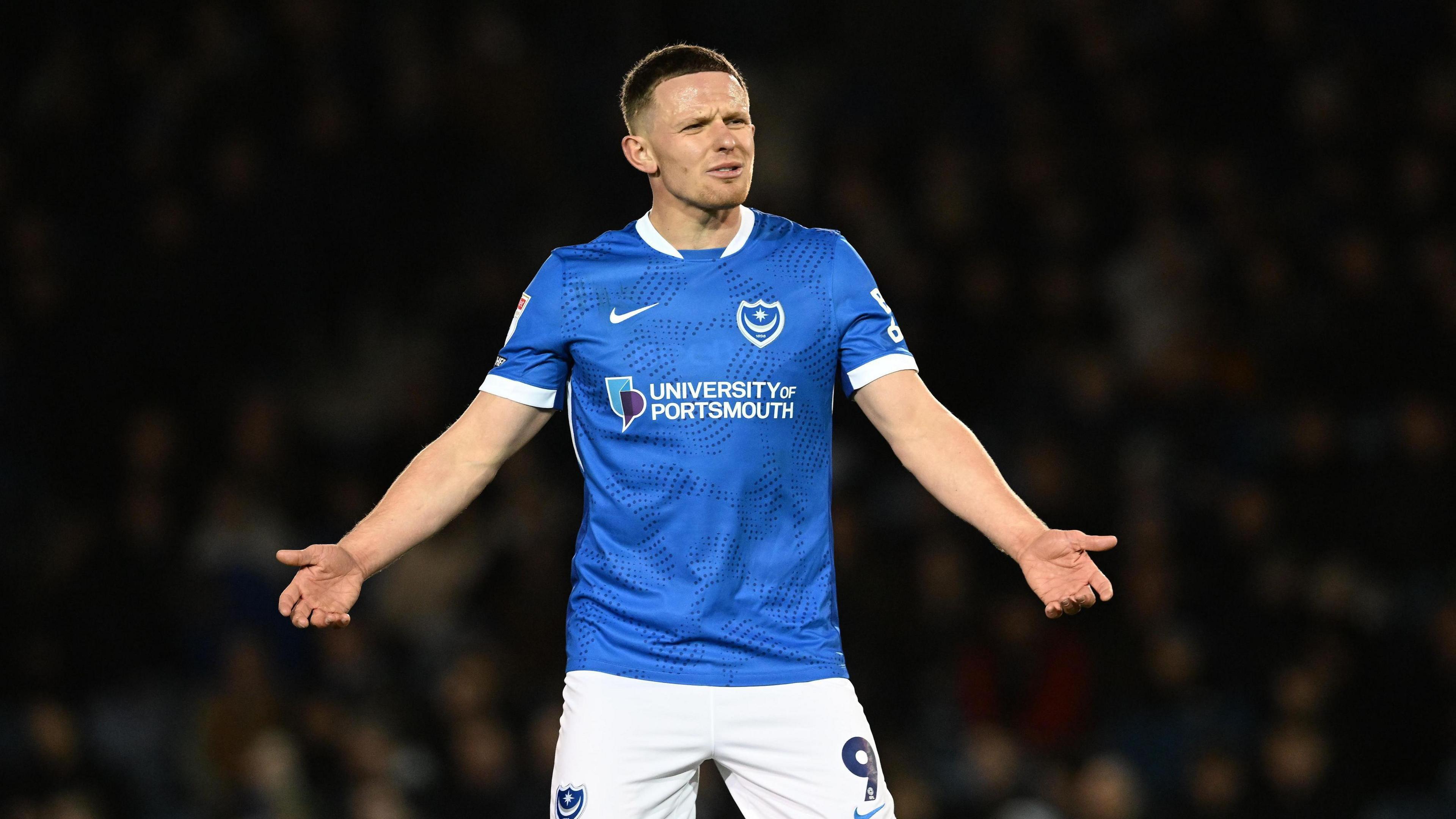 Colby Bishop holding out his arms in frustration while wearing a blue Portsmouth home kit and white shorts with the number 9 and the club badge printed on