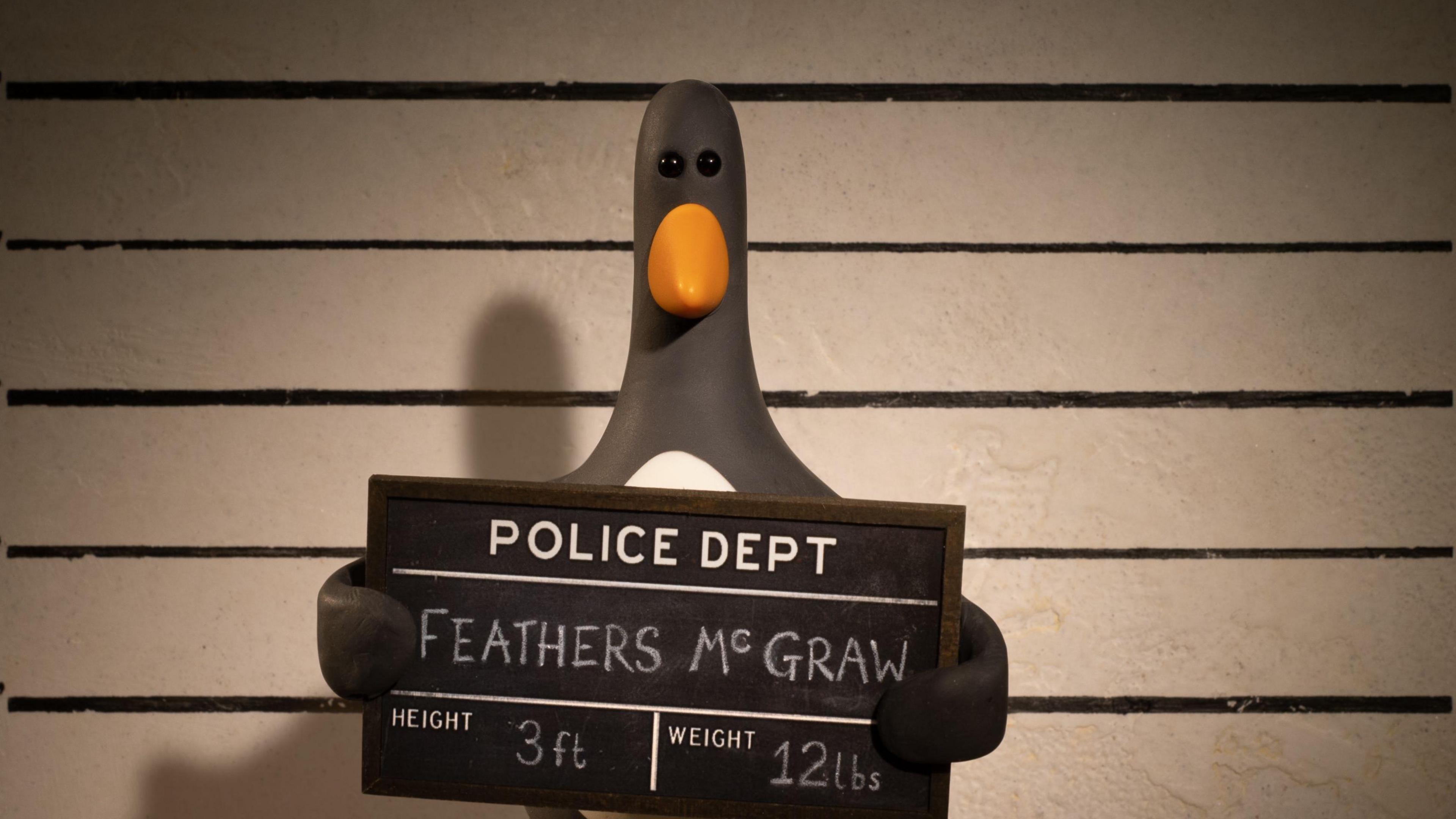 An animated version of a penguin stands in front of the camera in the style of a police mugshot, holding a borad which identifies him as Feathers McGraw and his height as 3ft and weight 12 pounds. 