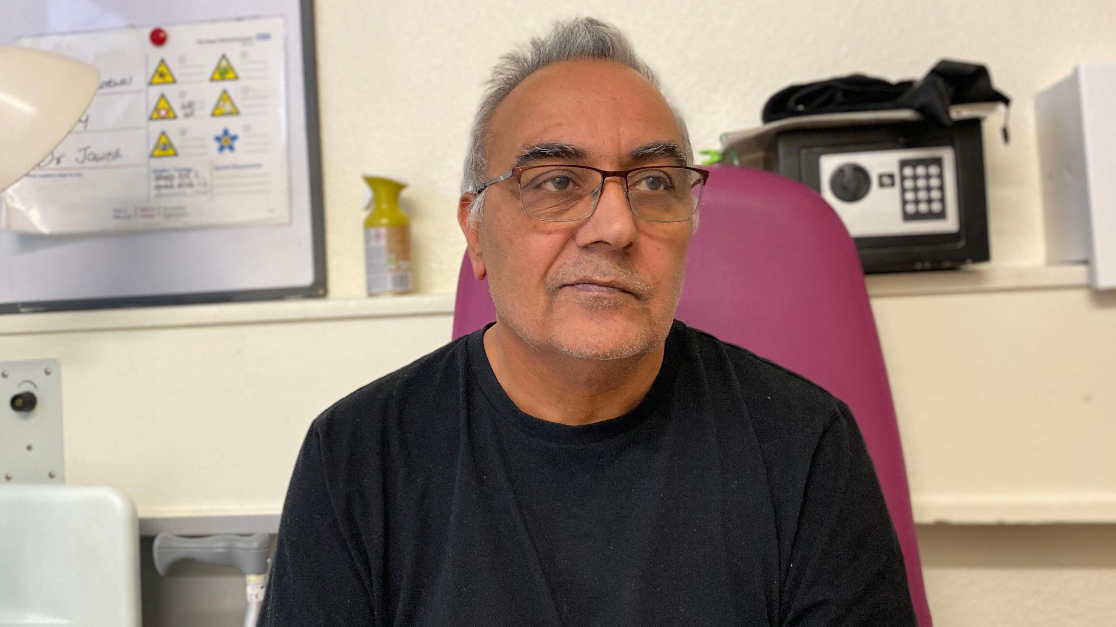 Ajay Purewal is wearing a black t-shirt and glasses. He is sitting on a pink chair in his hospital room.