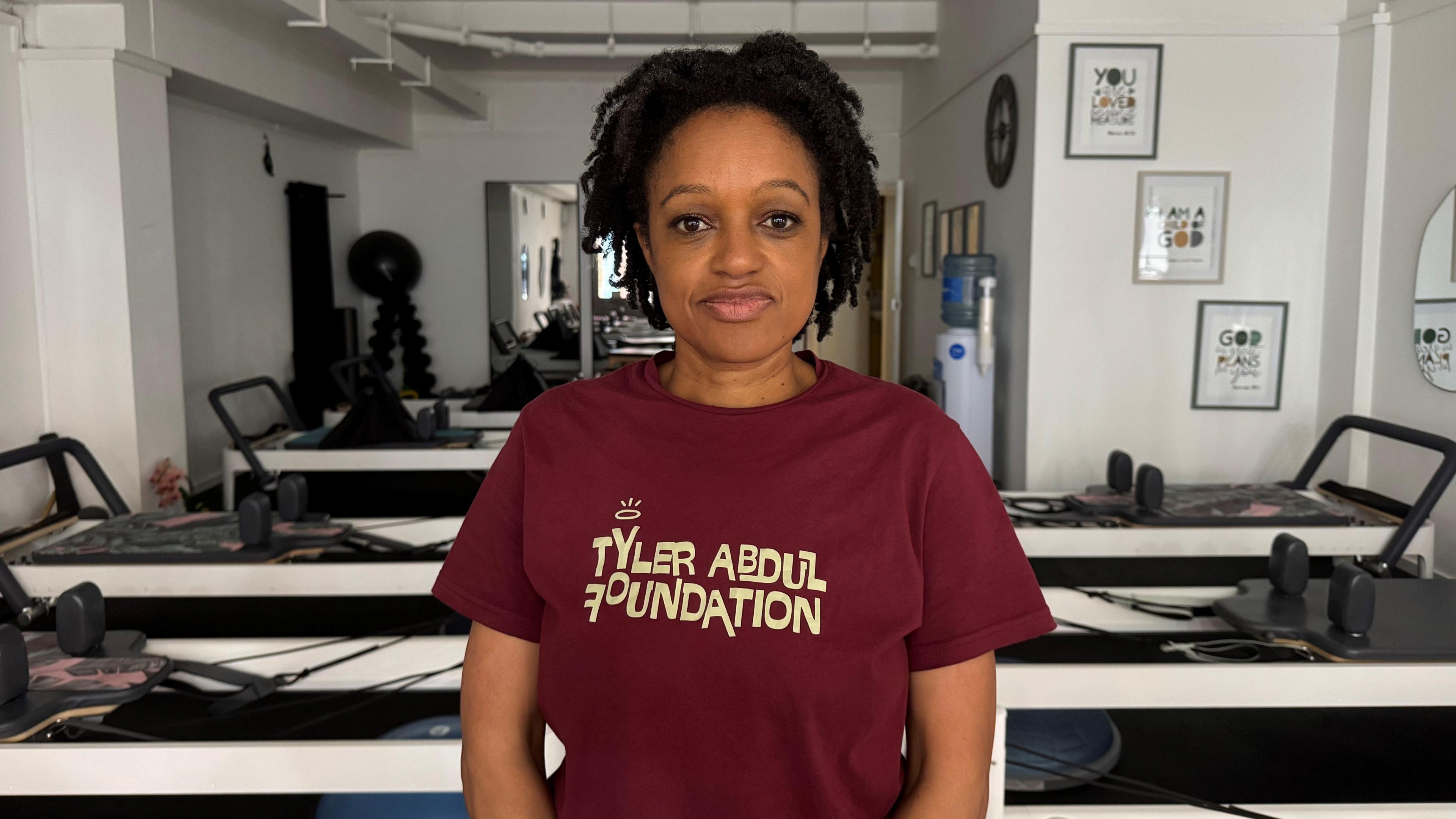 A woman wearing a burgundy shirt with cream writing on it, reading: "Tyler Abdul Foundation". She is standing in a Pilates studio, with equipment and a mirror in the background.