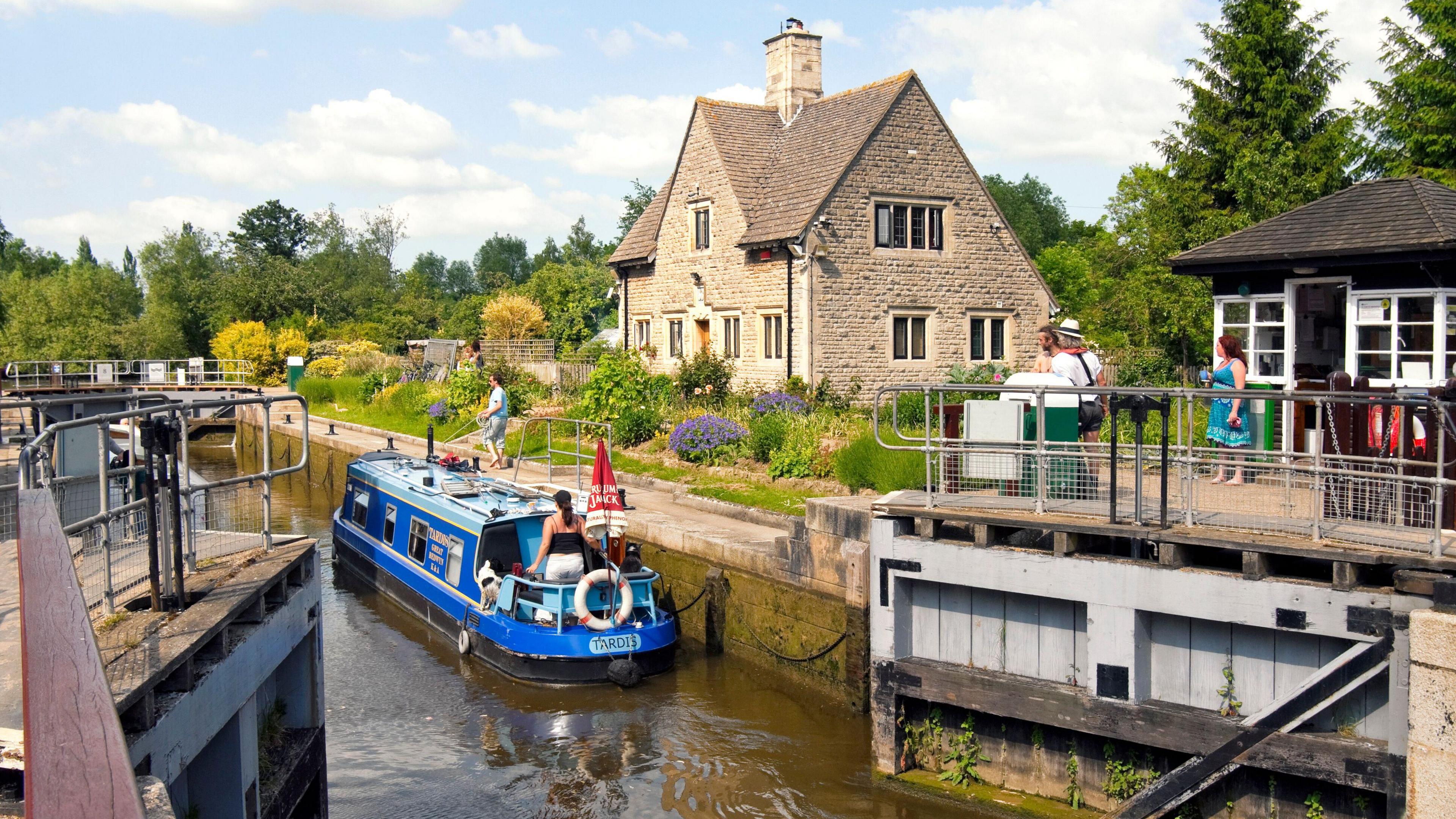 An open lock with a canal boat on the water with a person stood on it, on the background onland their is an old stone building and a wooden hut with three people stood outside