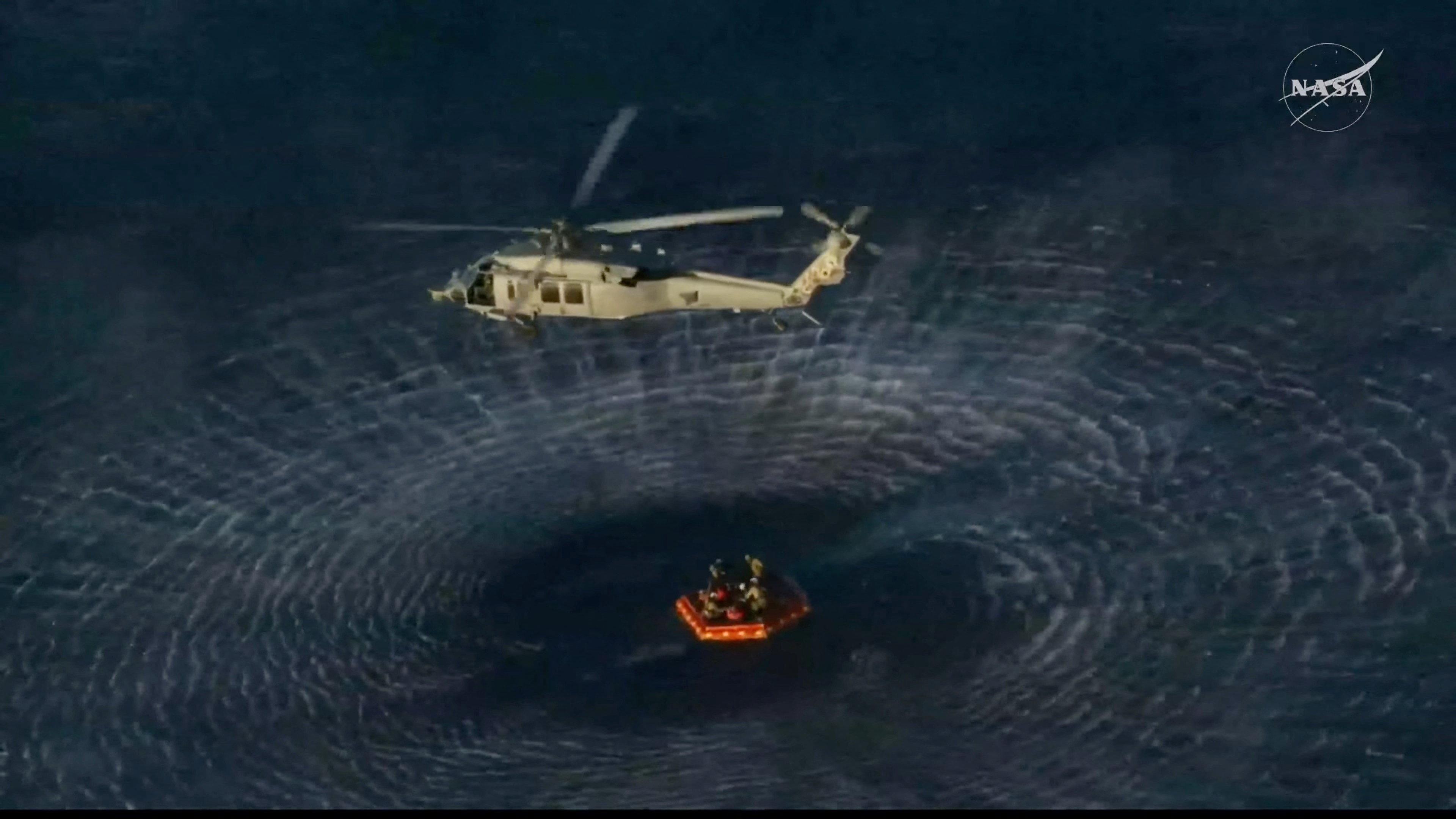 A helicopter whirs above an orange inflatable raft in the sea making outward ripples in the water.