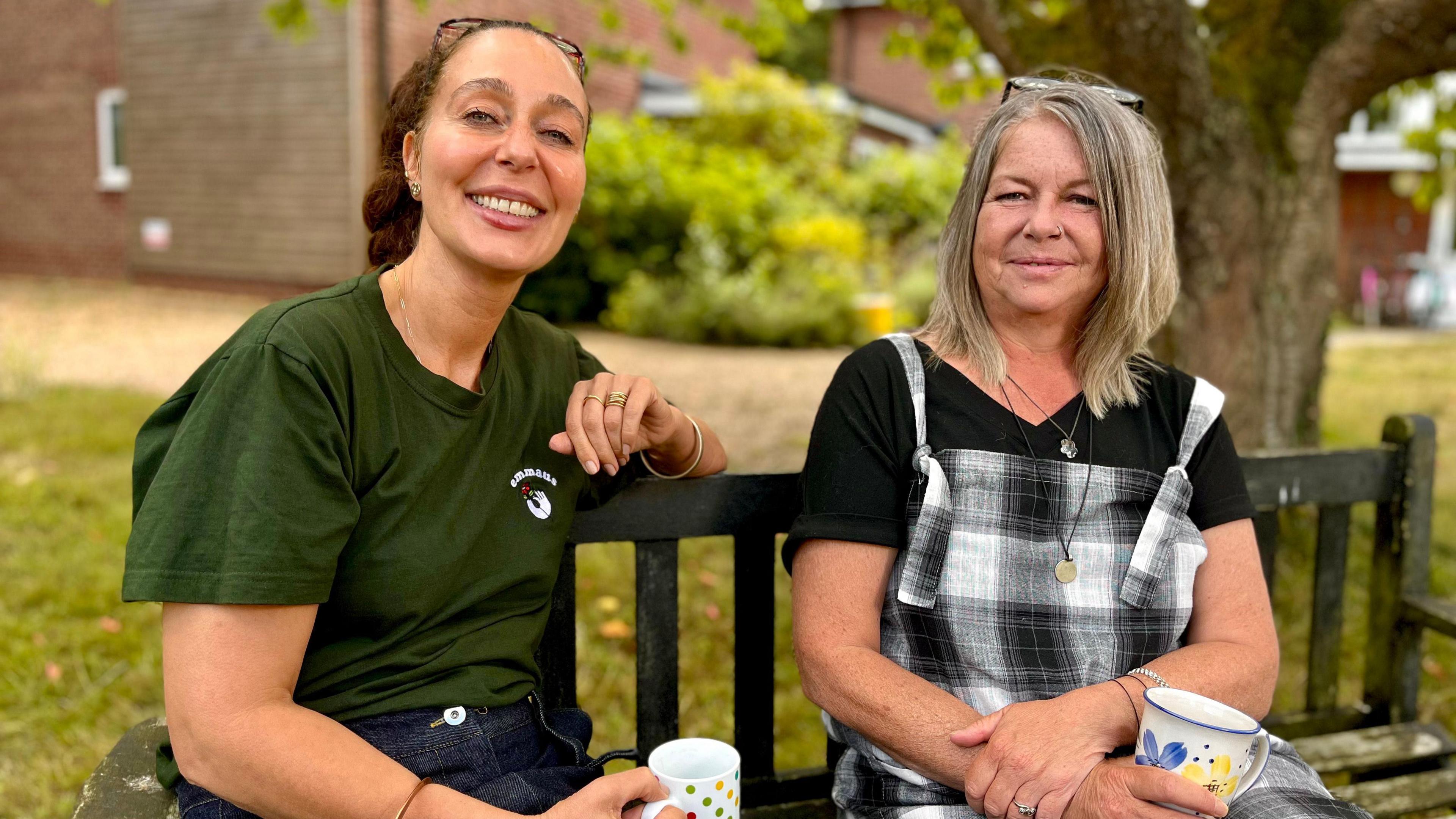 Two women sitting on a parking bench, both holding mugs of tea, with both looking to the camera. One has dark hair and is wearing a dark green T-shirt, while the other has blonde hair and is wearing black and white check dungarees over a black T-shirt.