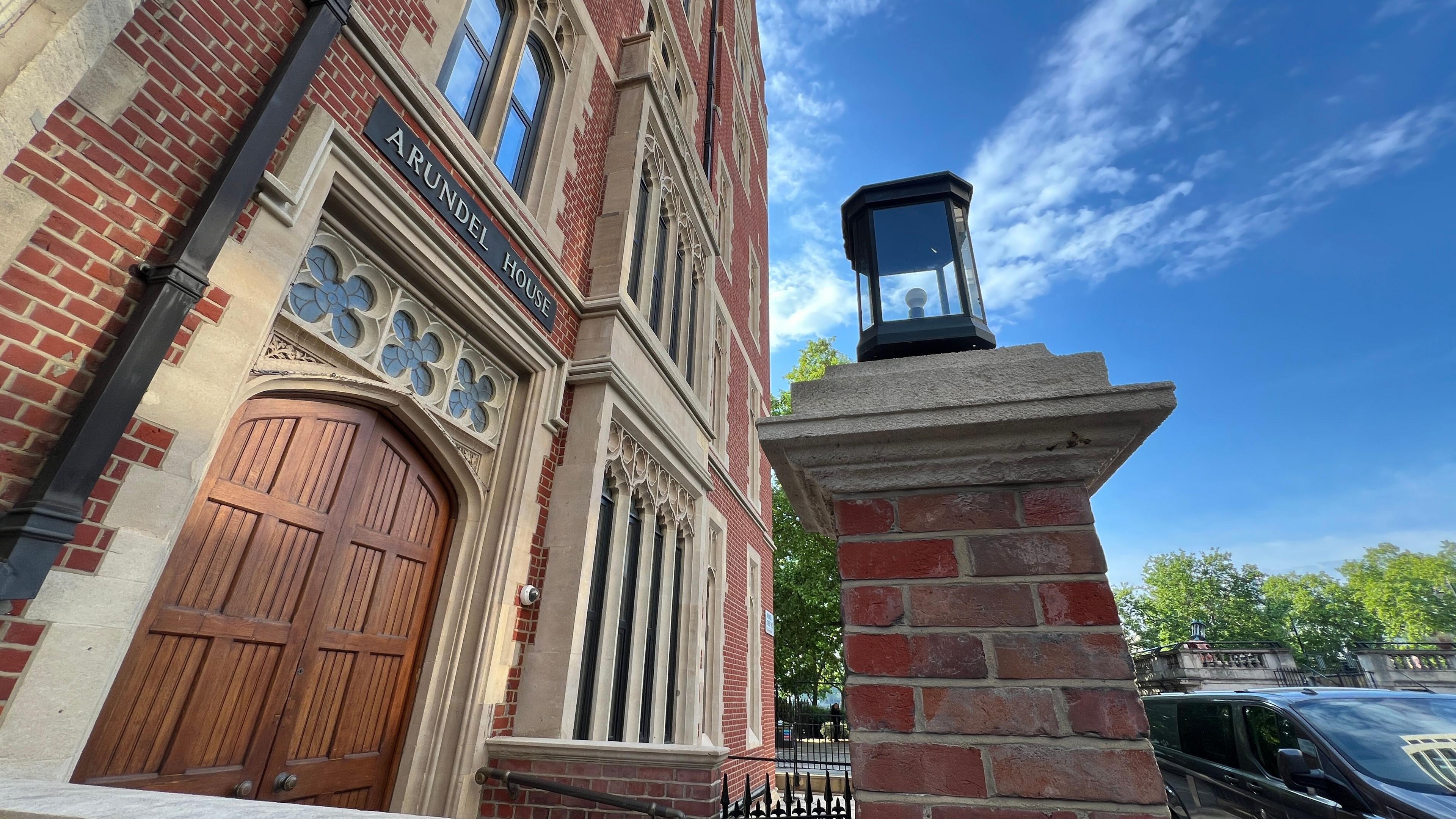 Arundel House is a red brick building with sandstone surrounding the doors and windows. There is a pair of closed wooden double doors at the front of the building with the words Arundel House on a black sign above the doorframe. There is a brick pillar with a black lantern on top at the front of the building, and there is a car parked next to the pillar. There are green trees in the distance and blue sky above.