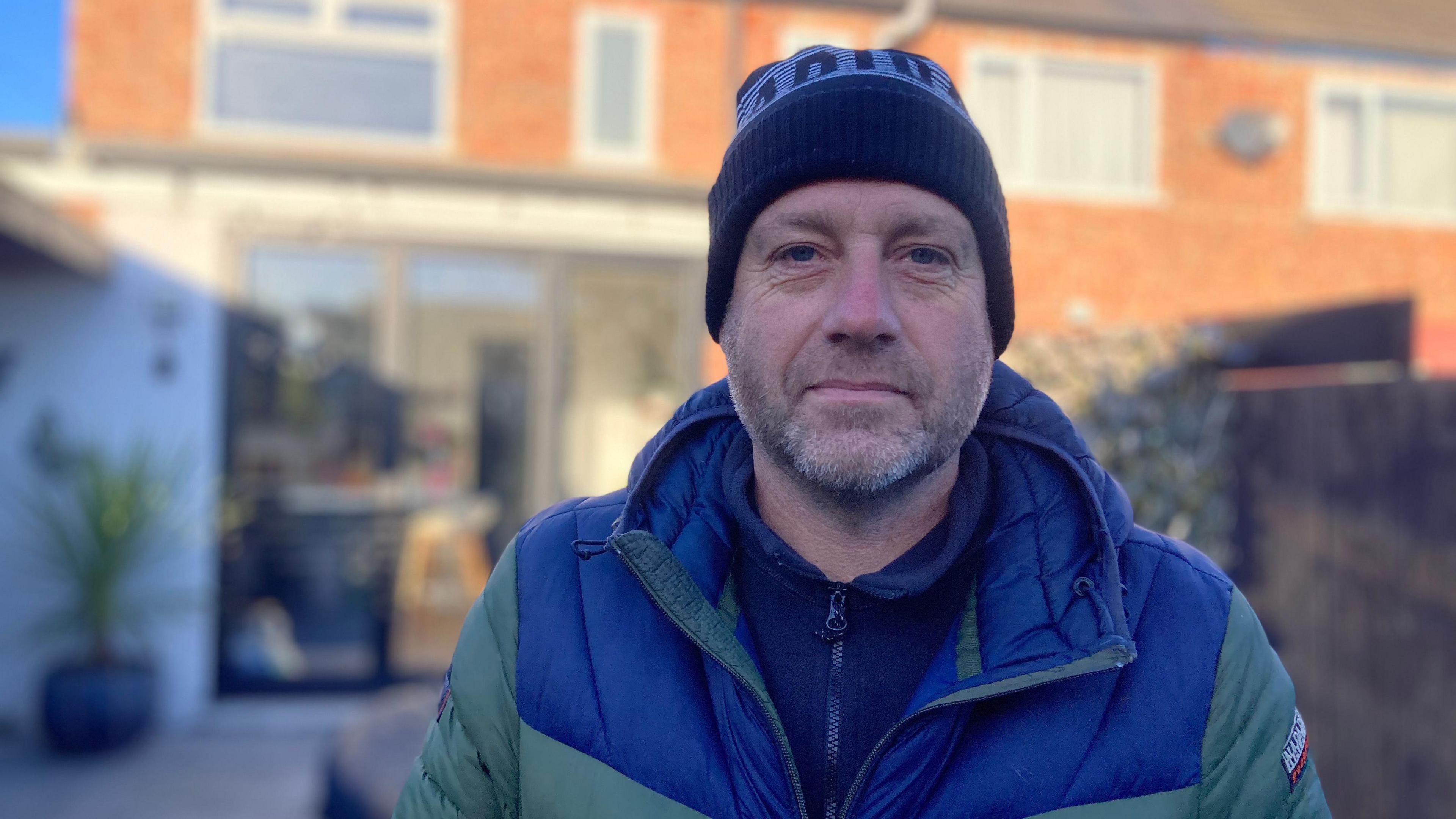 A head and shoulder shot of Lee Deller facing the camera. He is wearing a navy beanie hat and a blue and green padded coat with a navy zip-up jumper underneath. 