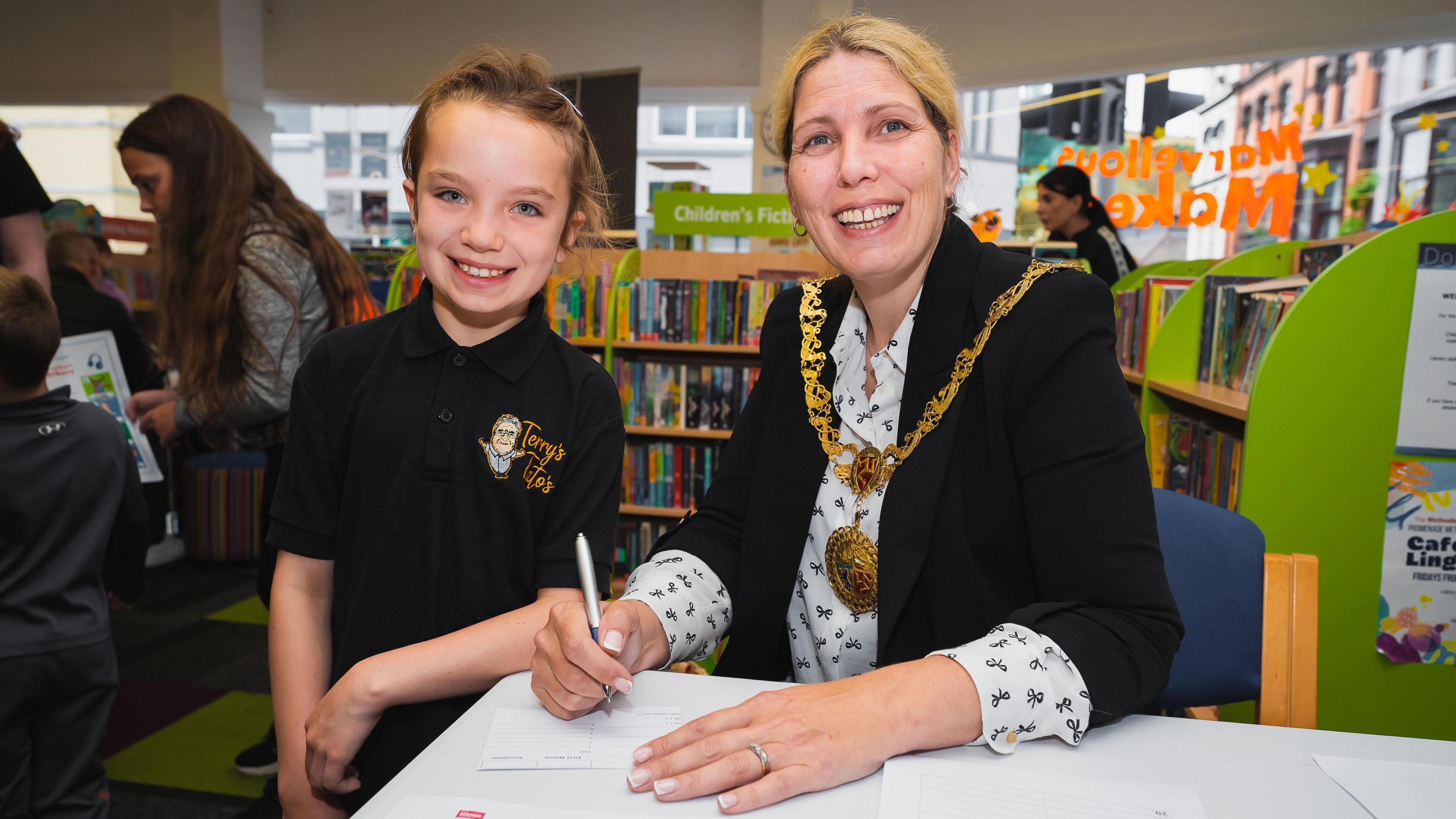 A primary school girl with blue eyes stood next to a woman sat down, the mayor of Douglas wearing an extravagant gold chain that connects to a cape, in a library.