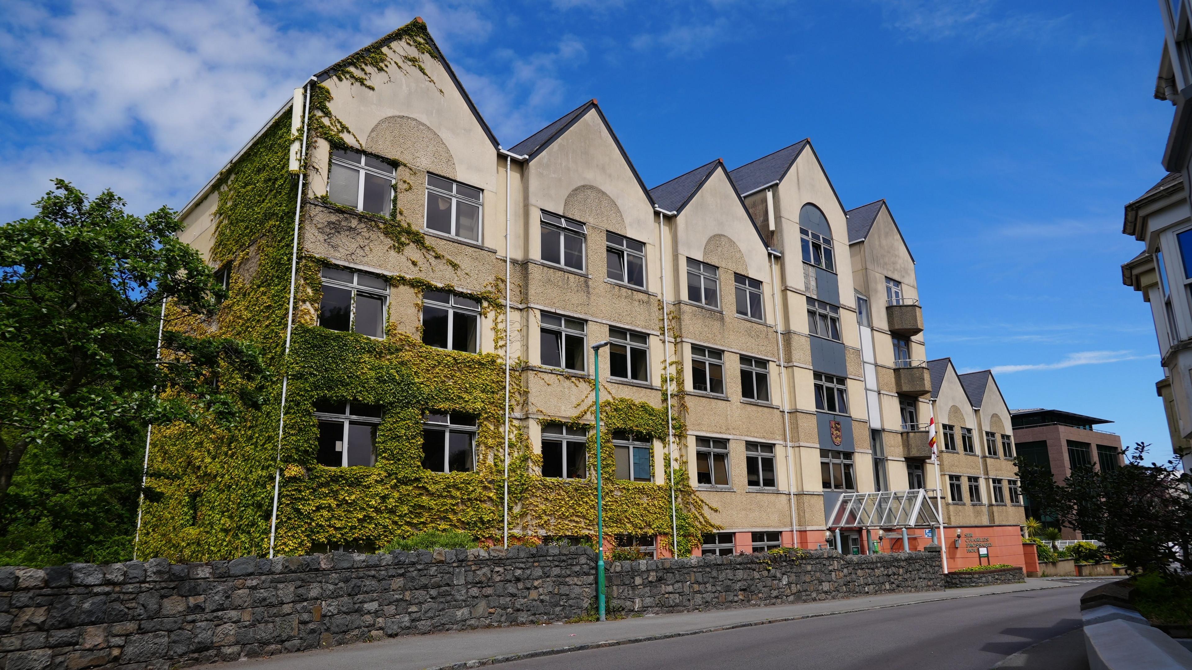 A yellow building wikth six triangular pointed roofs. The lower left third of the building is covered in green ivy. 