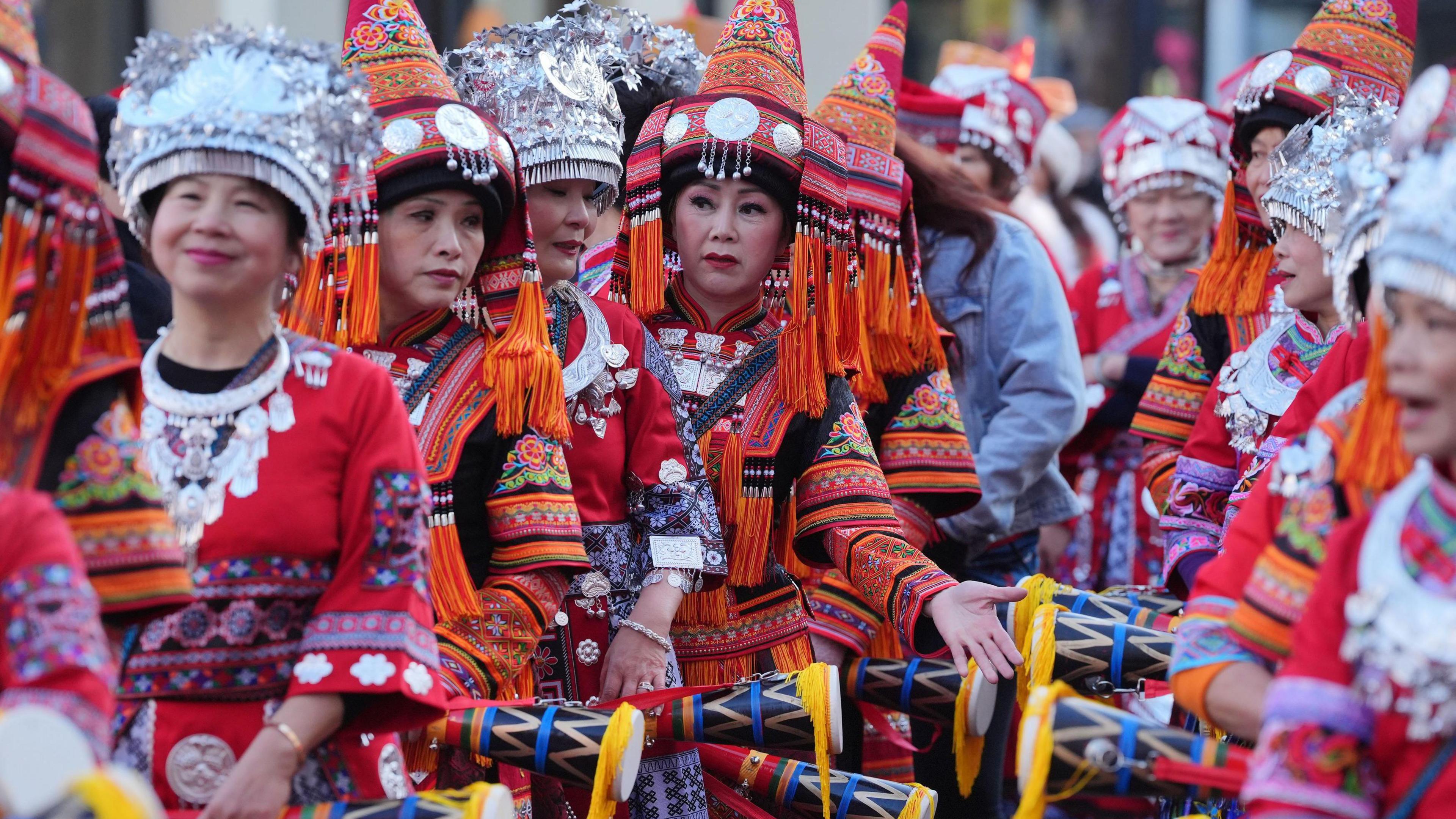 A group of women wearing colourful, traditional Asian clothing, including silver headdresses and red embroidered outfits, stand in a row holding decorated drumsticks.