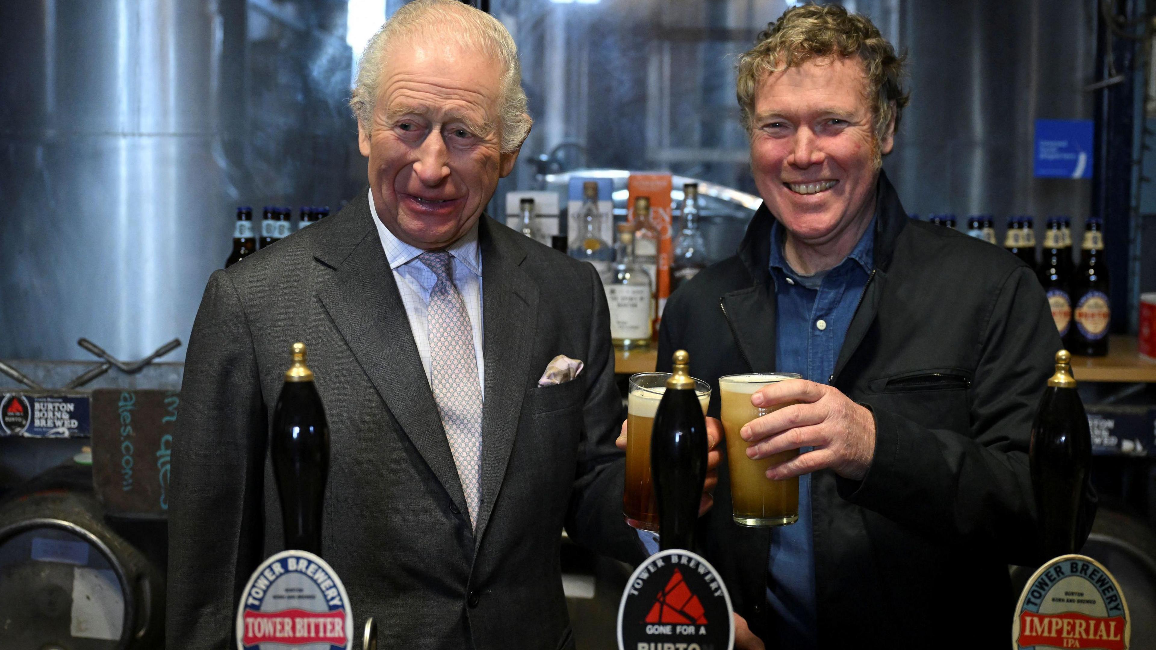 King Charles smiles as he holds a pint behind a bar alongside brewery owner John Mills, who is also holding a pint.