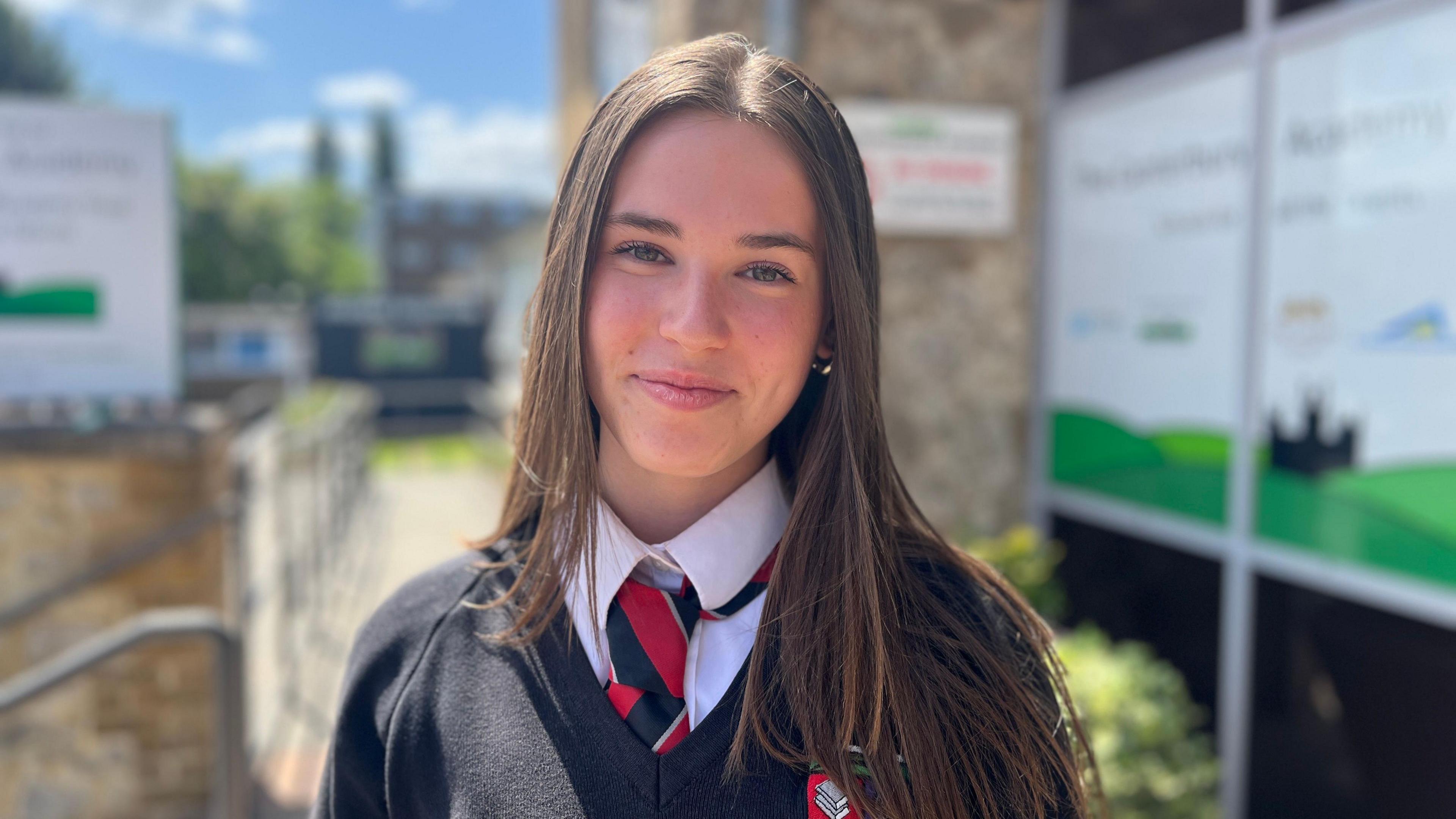 Teenager Molly with long brown hair smiles to the camera. She's wearing school uniform including a black and red tie.