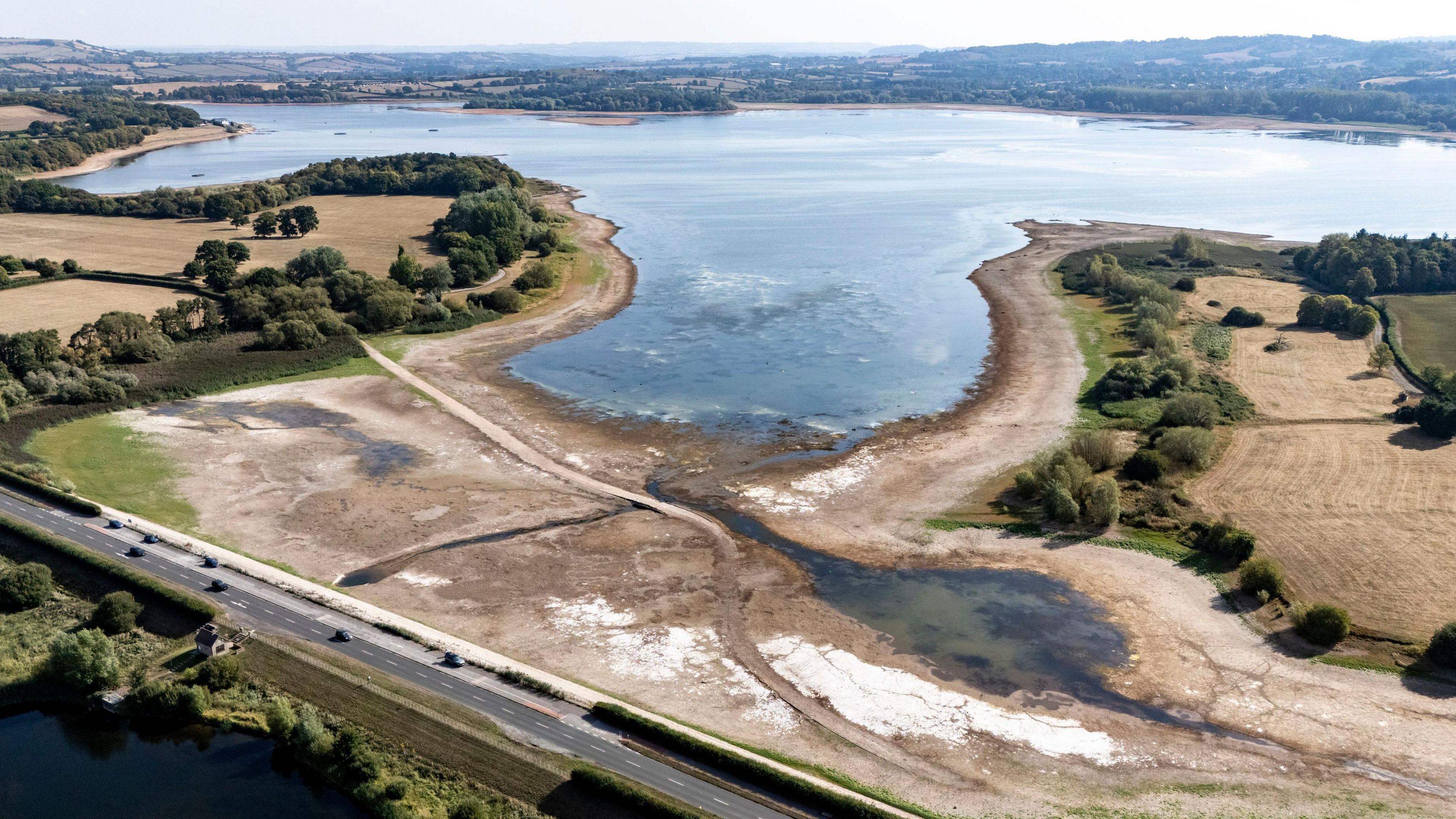 An aerial shot of a lake which is receding from its bank, leaving dry and brown ground behind. The grass fields around the lake are brown as well, with a few green trees dotted around.