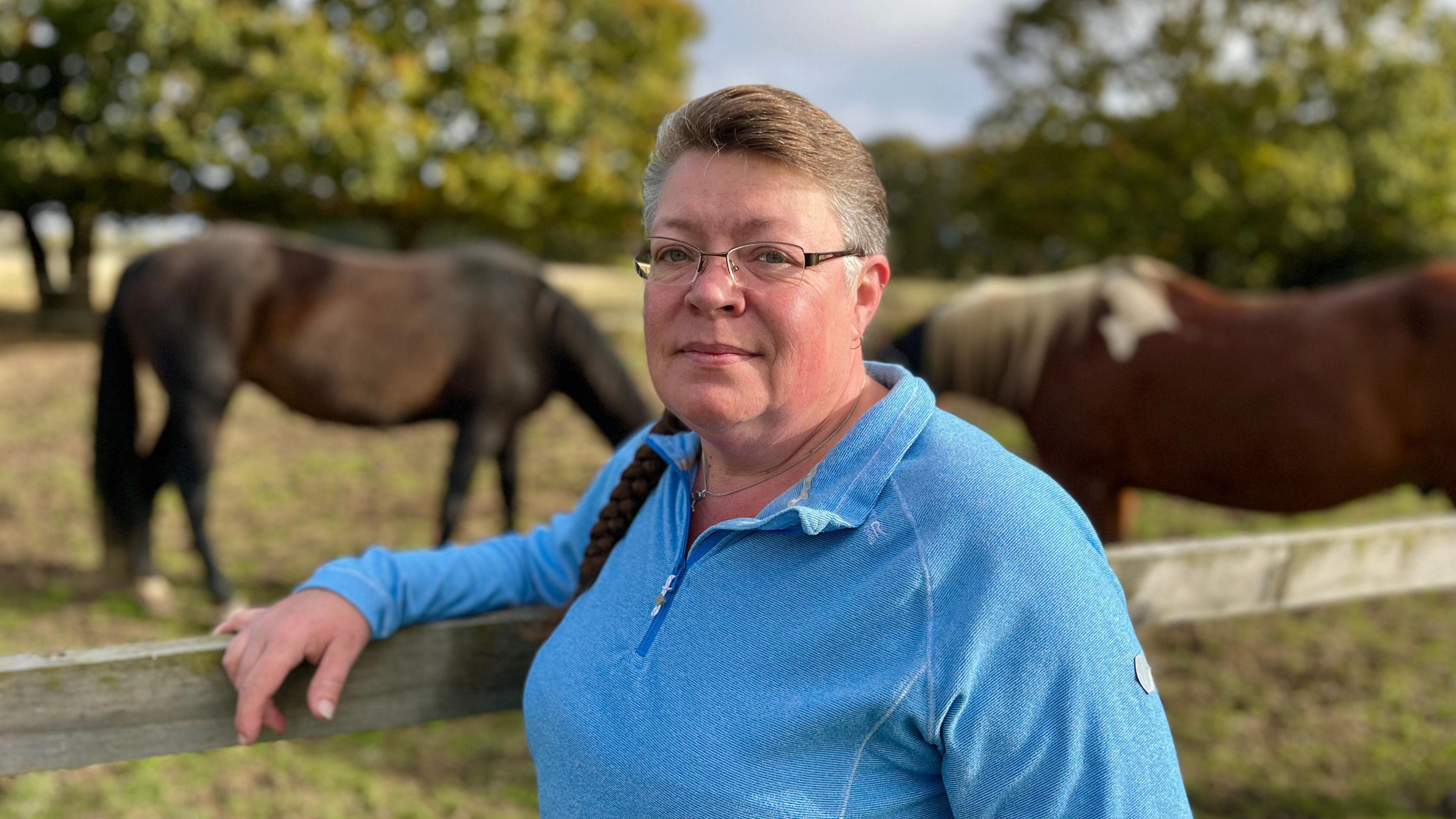 Rachel Kennedy looks directly at the camera, she is photographed outside wearing a blue jumper with her arm resting on a wooden fence. Two horses are in the background in a field.