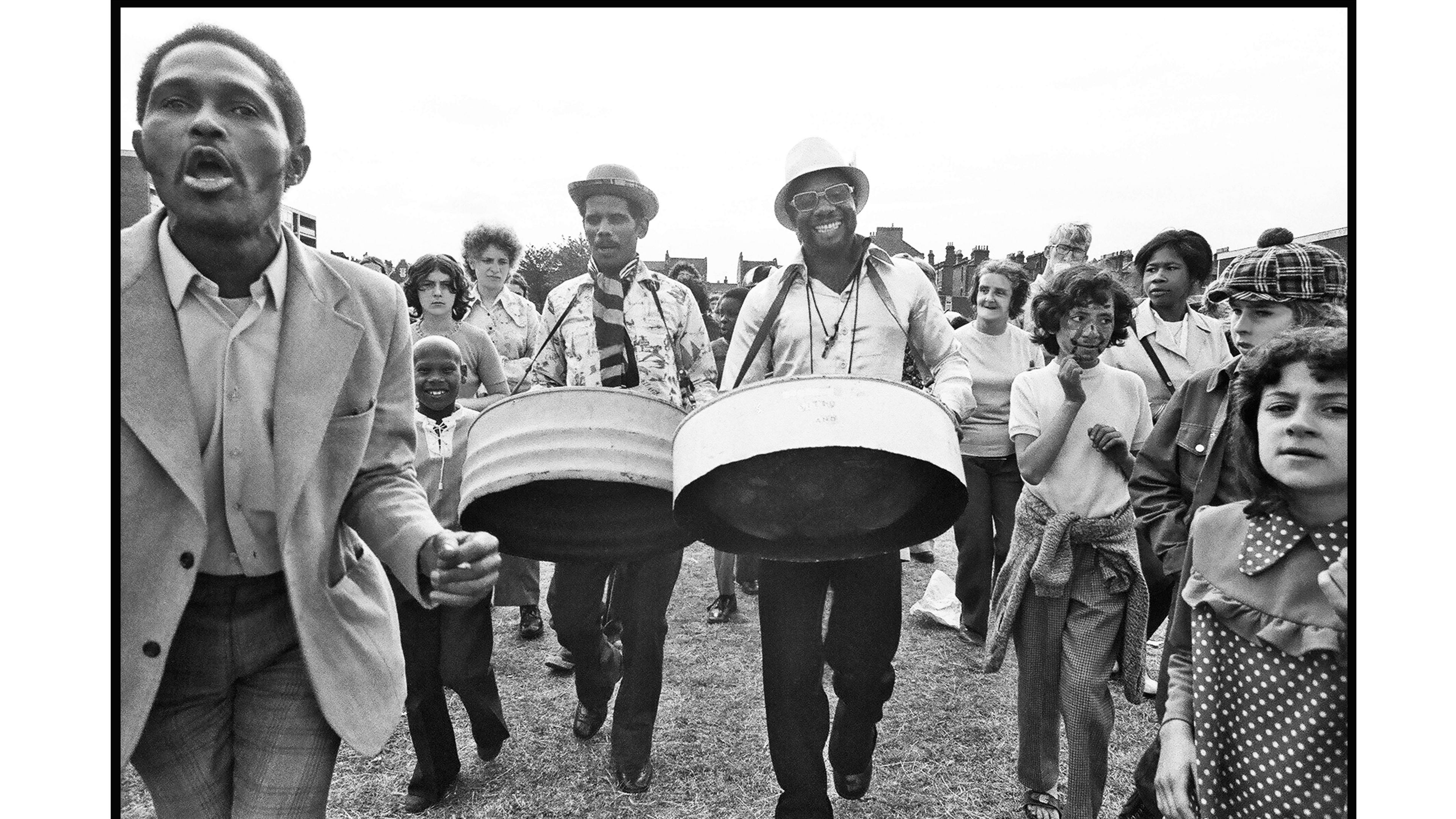 A black and white image of two steel drum players surrounded by children. 