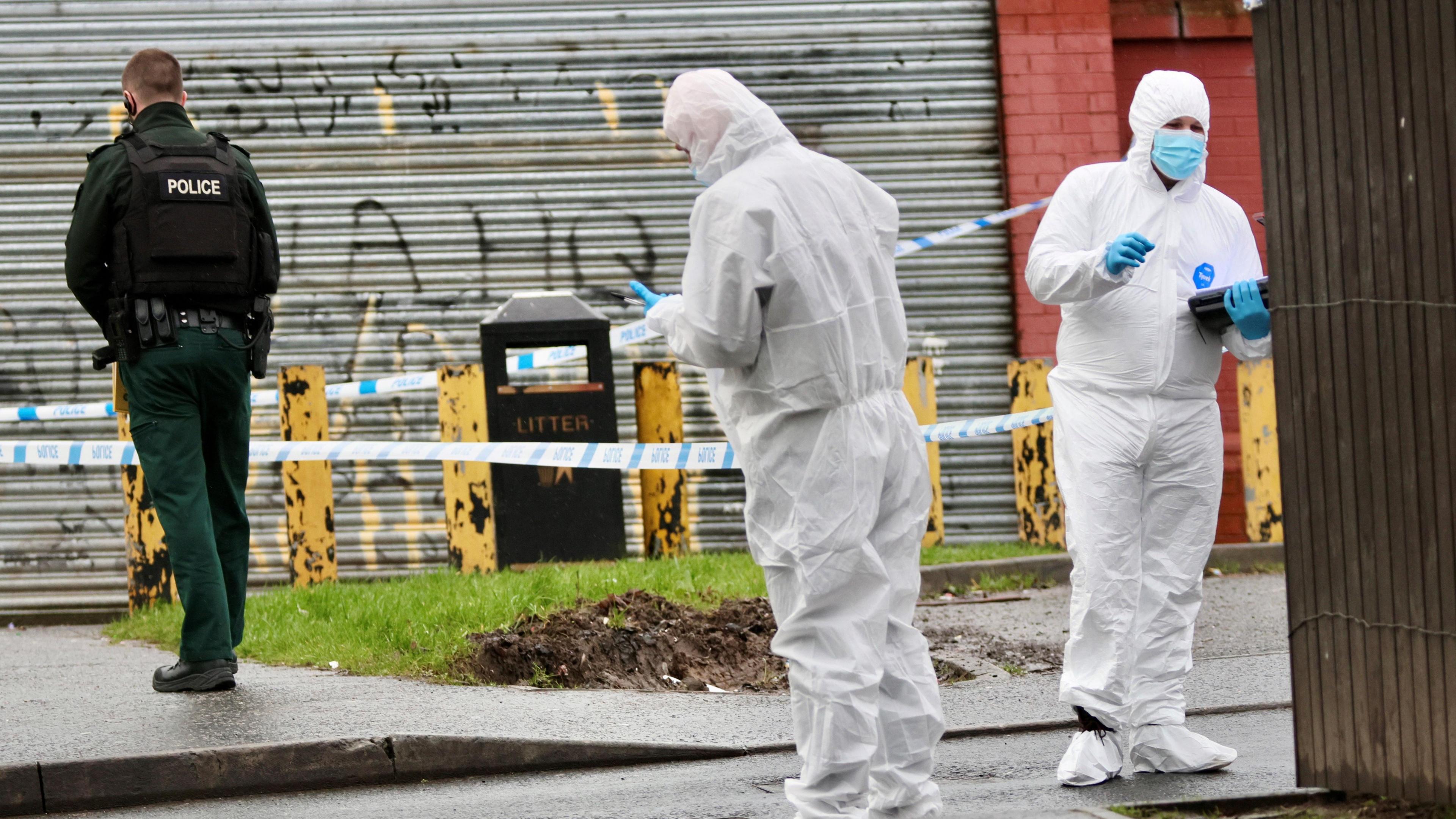 Two people in white forensic suits standing on a street near a police cordon. A Uniformed officer is walking away from the camera. In the background a premises with its shutter down and a bin that says "litter" on it.