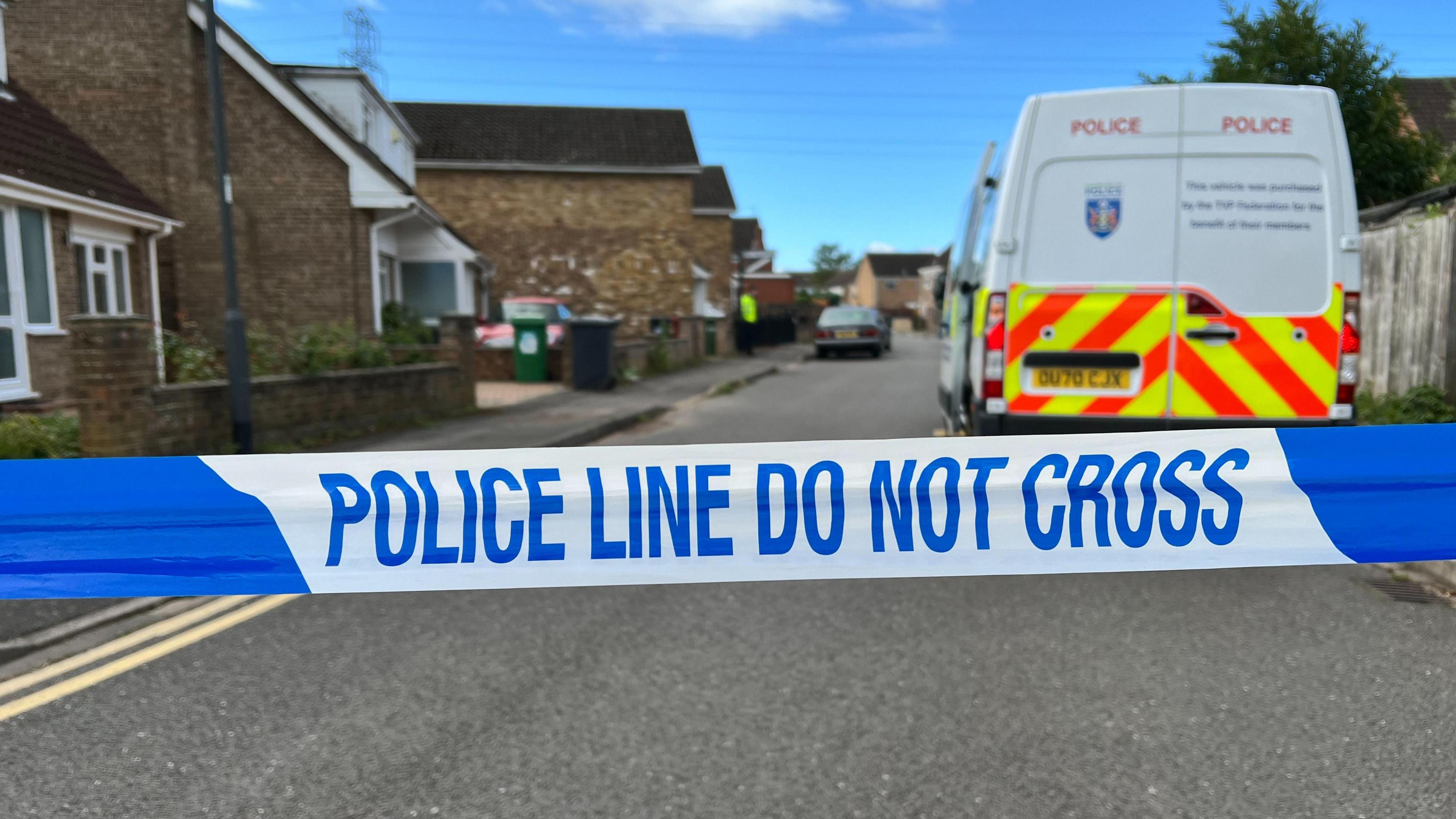 A line of blue ad white police tape saying 'police line do not cross' with a police van and a residential street in the background under a blue sky.