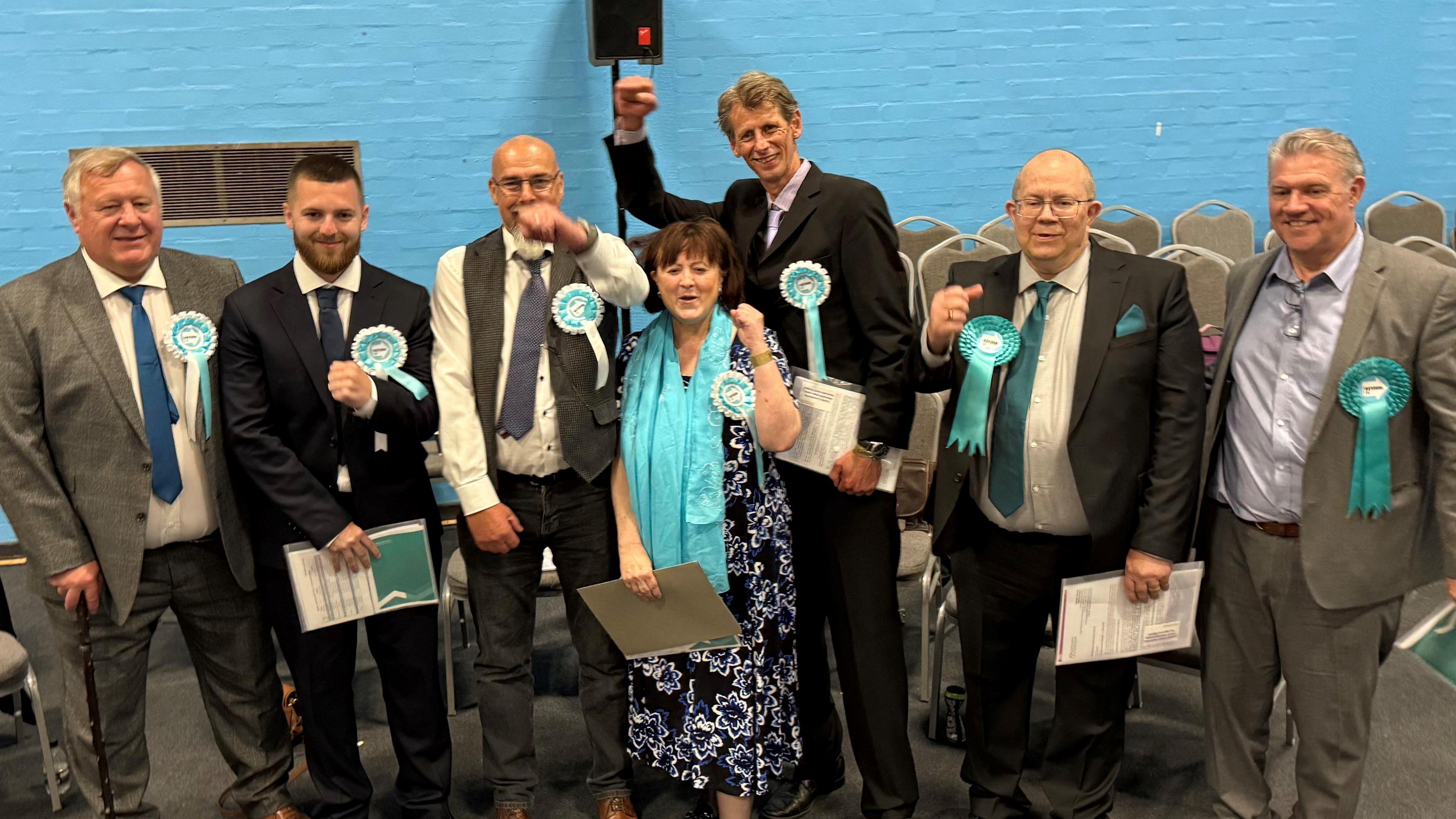 Seven Reform UK politicians are seen smiling and celebrating. They are wearing light blue rosettes and are stood against a blue wall.