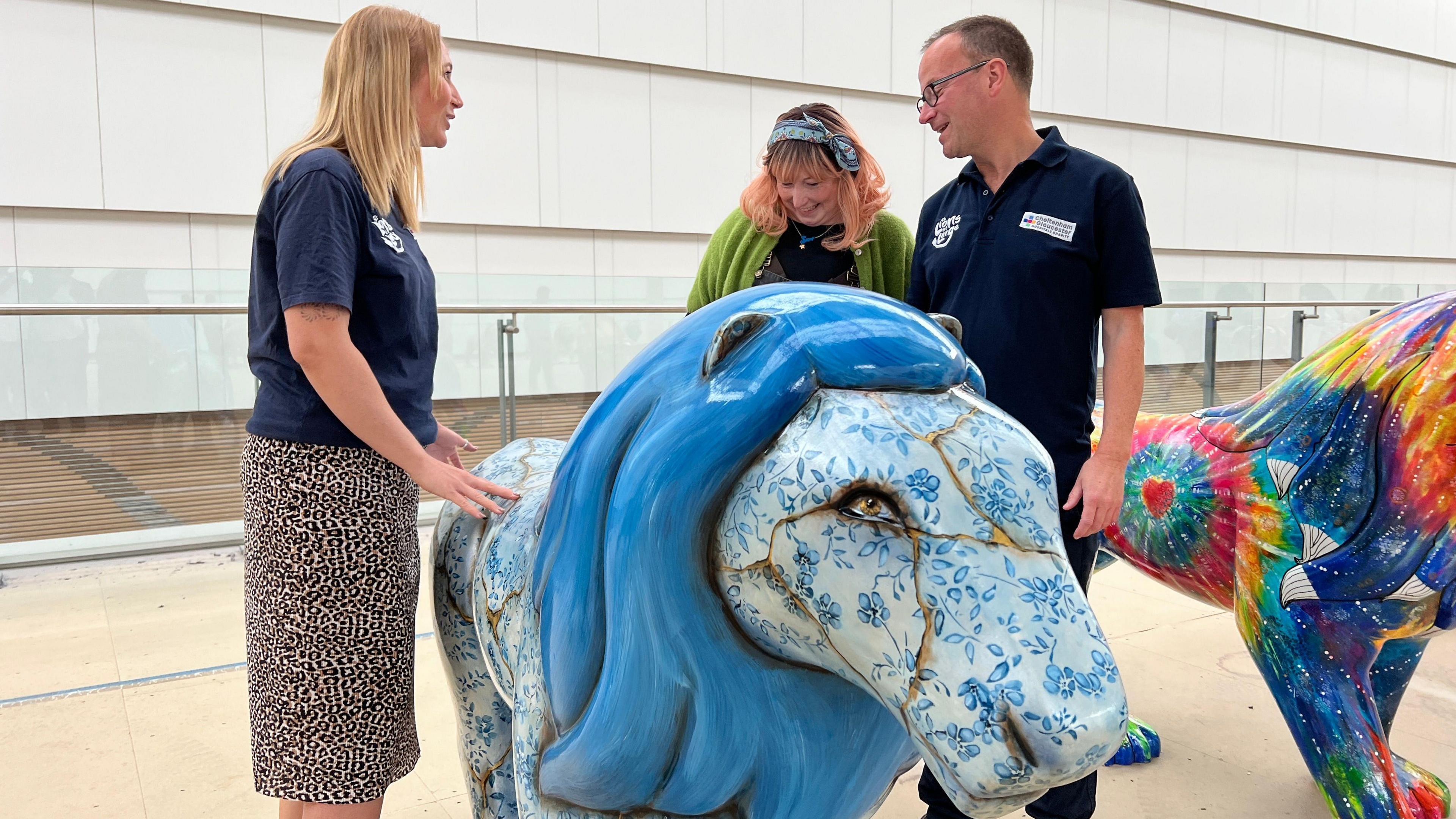 Sam looks down at her lion with two other people wearing branded tops. Behind them is a brightly coloured lion covered with a tie-dye print.