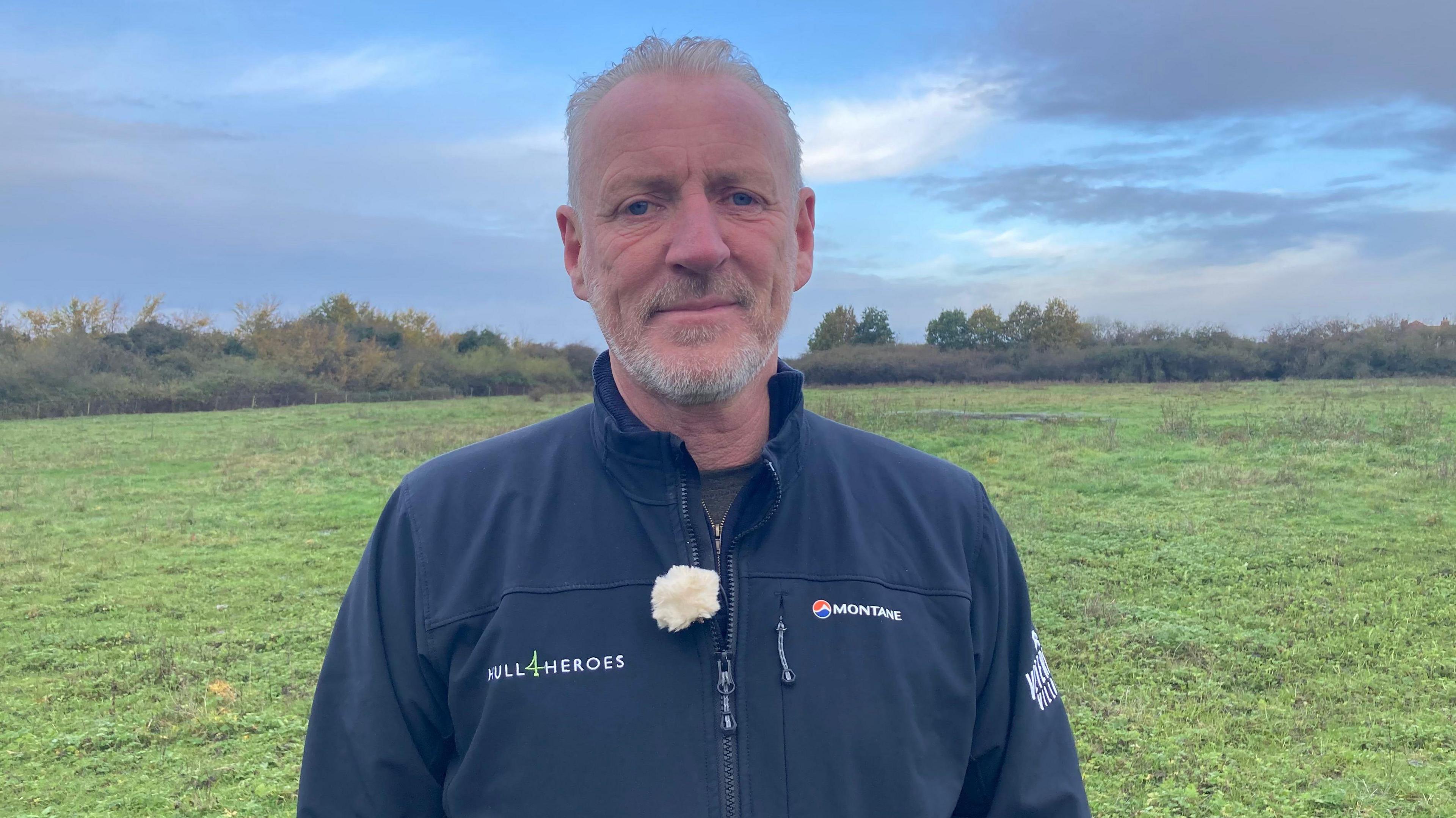 Paul Matson, who has short grey hair and a short grey beard, stands in a grassy field. He is wearing a blue jacket with the Hull 4 Heroes logo on.