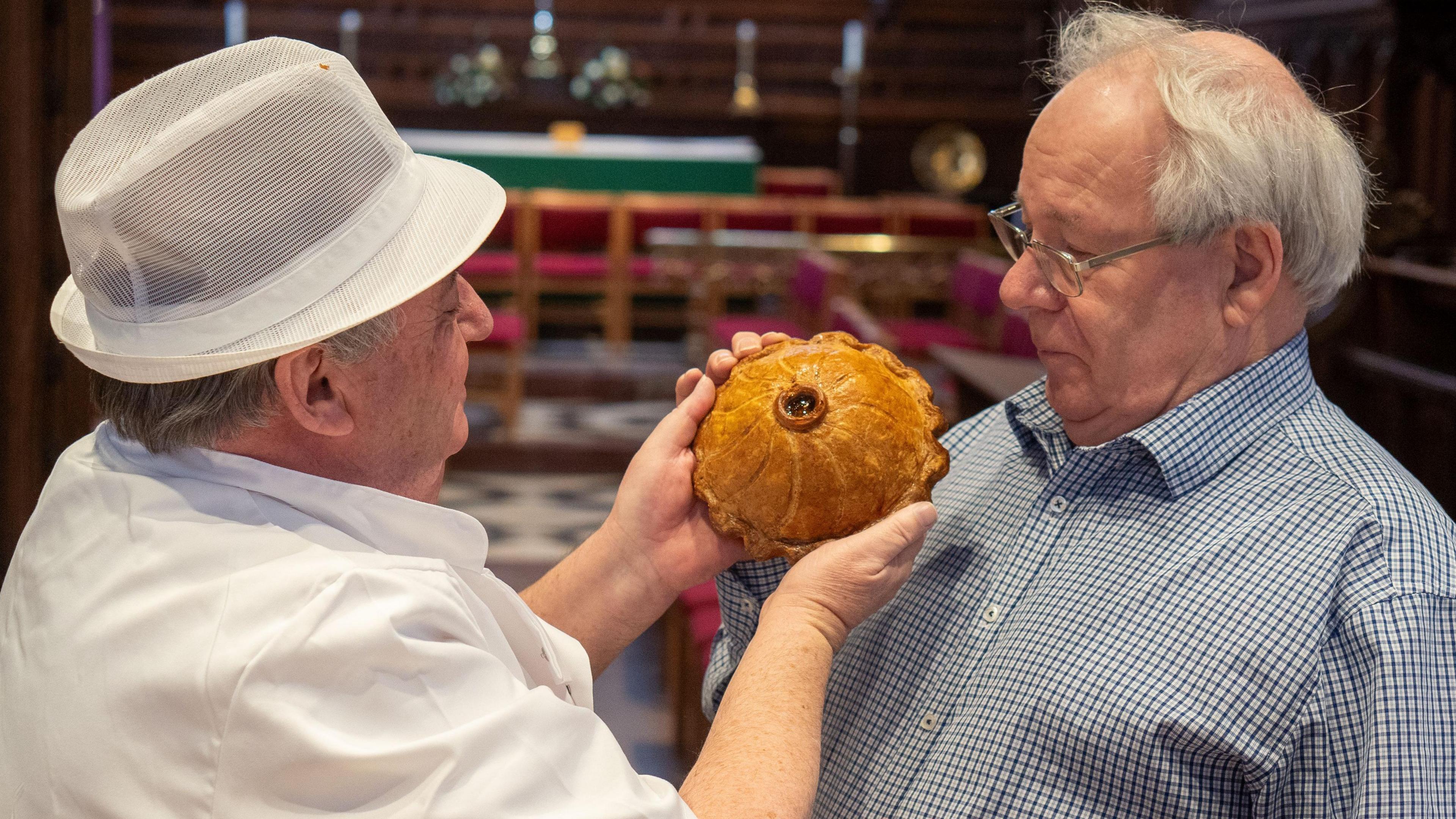 Mike Holling is wearing a white uniform and white hat. He is holding a pie up to the face of Matthew who is inspecting it. Matthew is wearing glasses and has white hair.