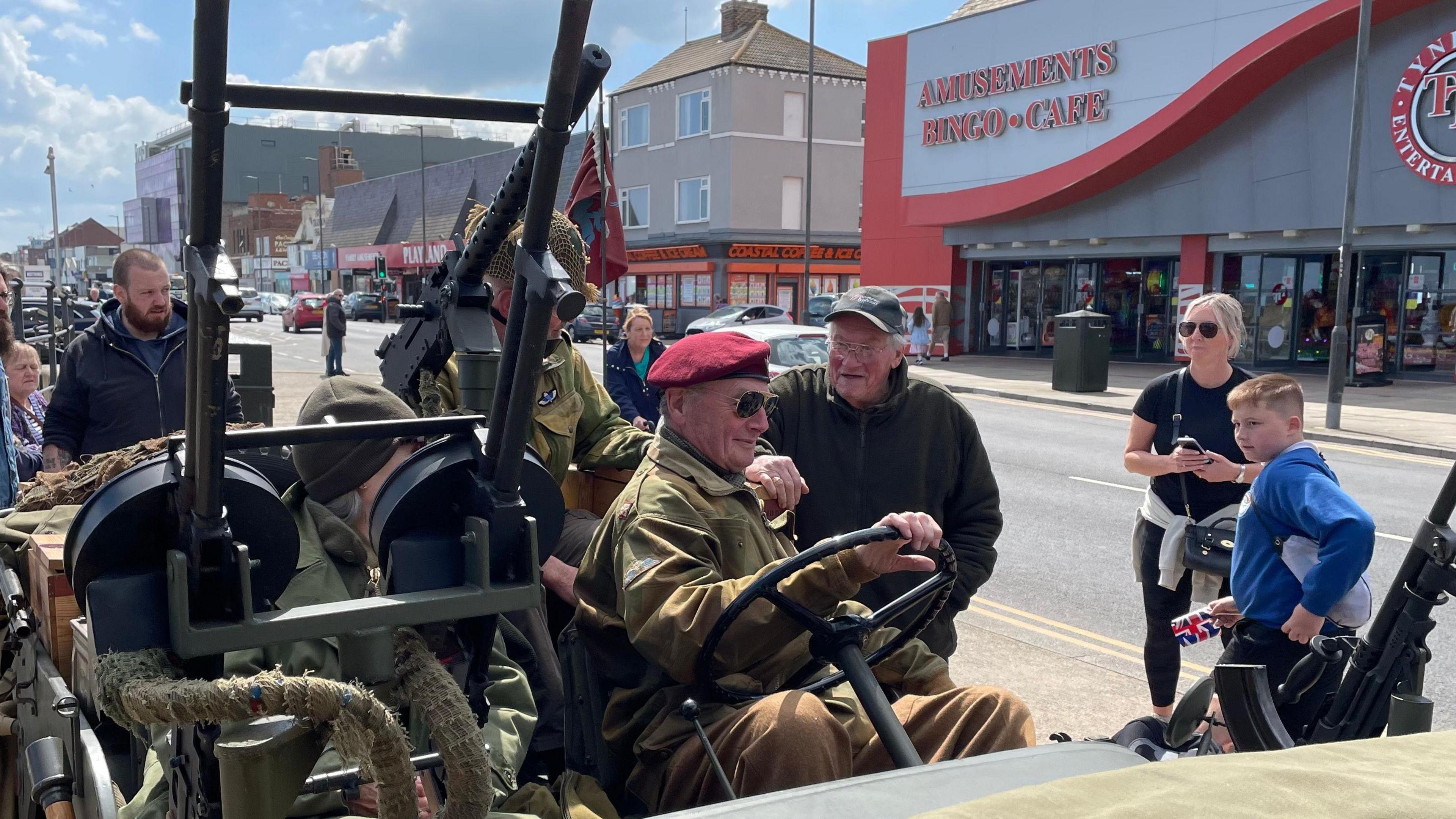 A man in a red beret at the wheel of a military vehicle, with others observing