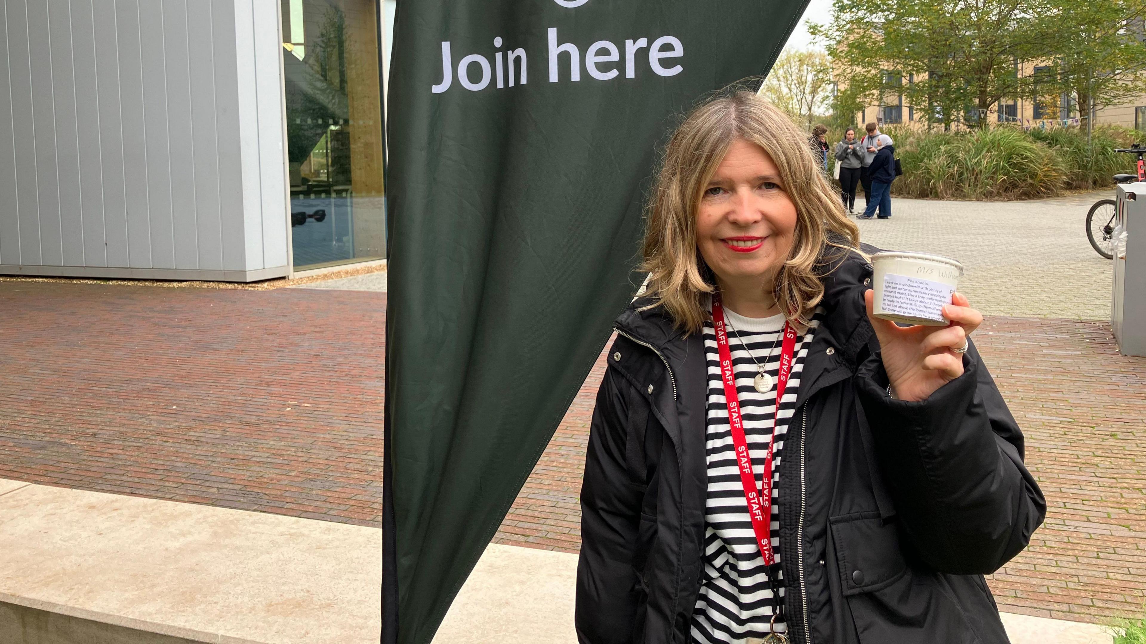 Jo Marten, who has blonde hair and is wearing a black coat and a white-and-black striped top holds up a pot of freshly planted pea seeds.