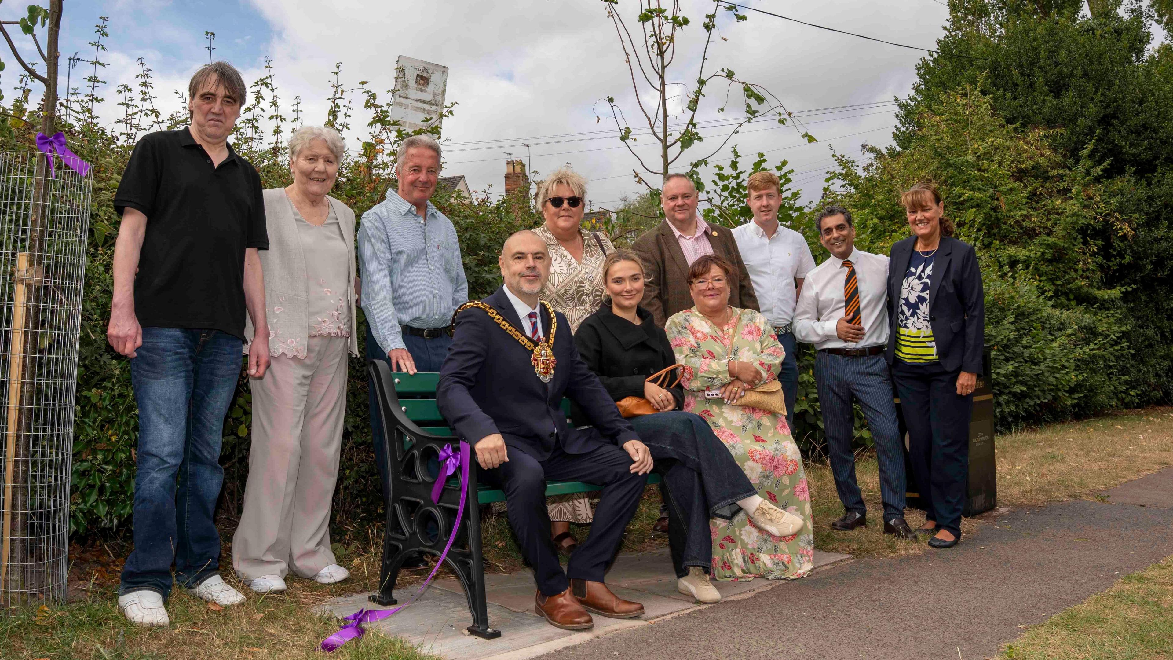Wolverhampton mayor Craig Collinswood is sitting on the memorial bench. He is wearing his chain of office and a dark suit. He is surrounded by a group of people who are the friends and family of former councillor Paddy Bradley