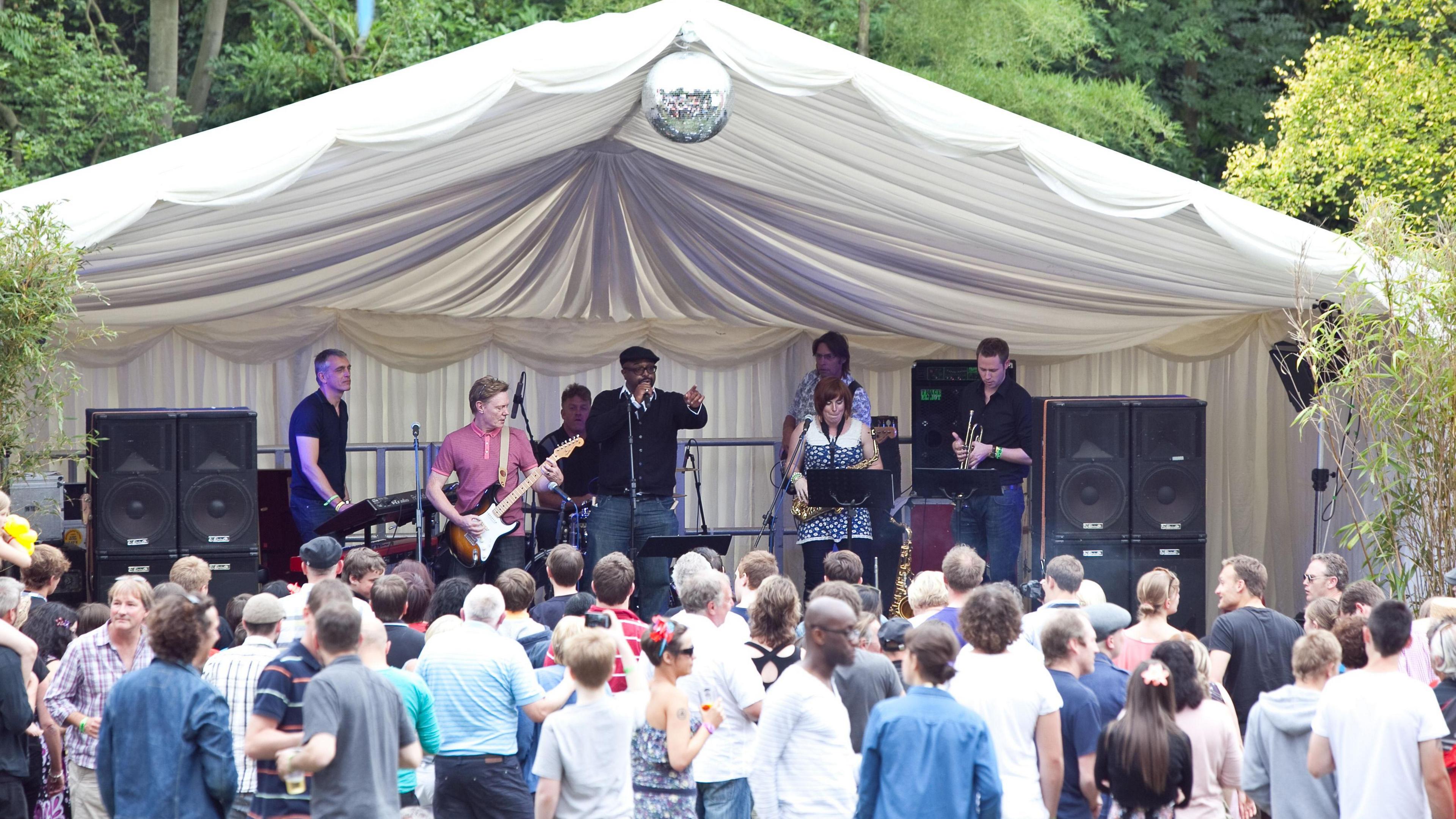 Crowds are watching as a group of men and women play instruments on a stage. There is a disco ball above their heads and trees in the background.