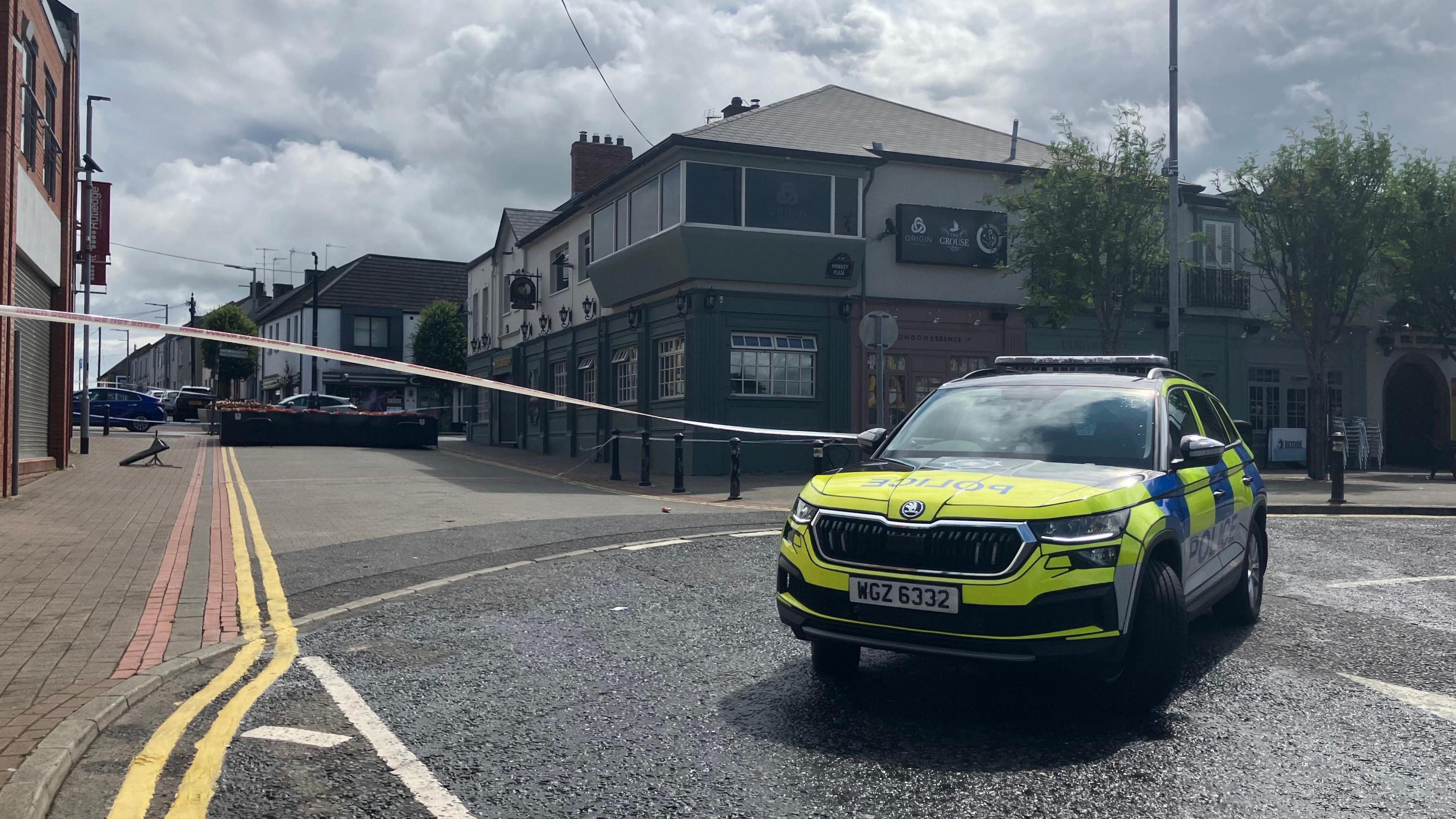 A marked police car is in the foreground. White police tape is across the street. In the background you can see buildings on either side of the street and double yellow lines along the left hand side as far as the eye can see.