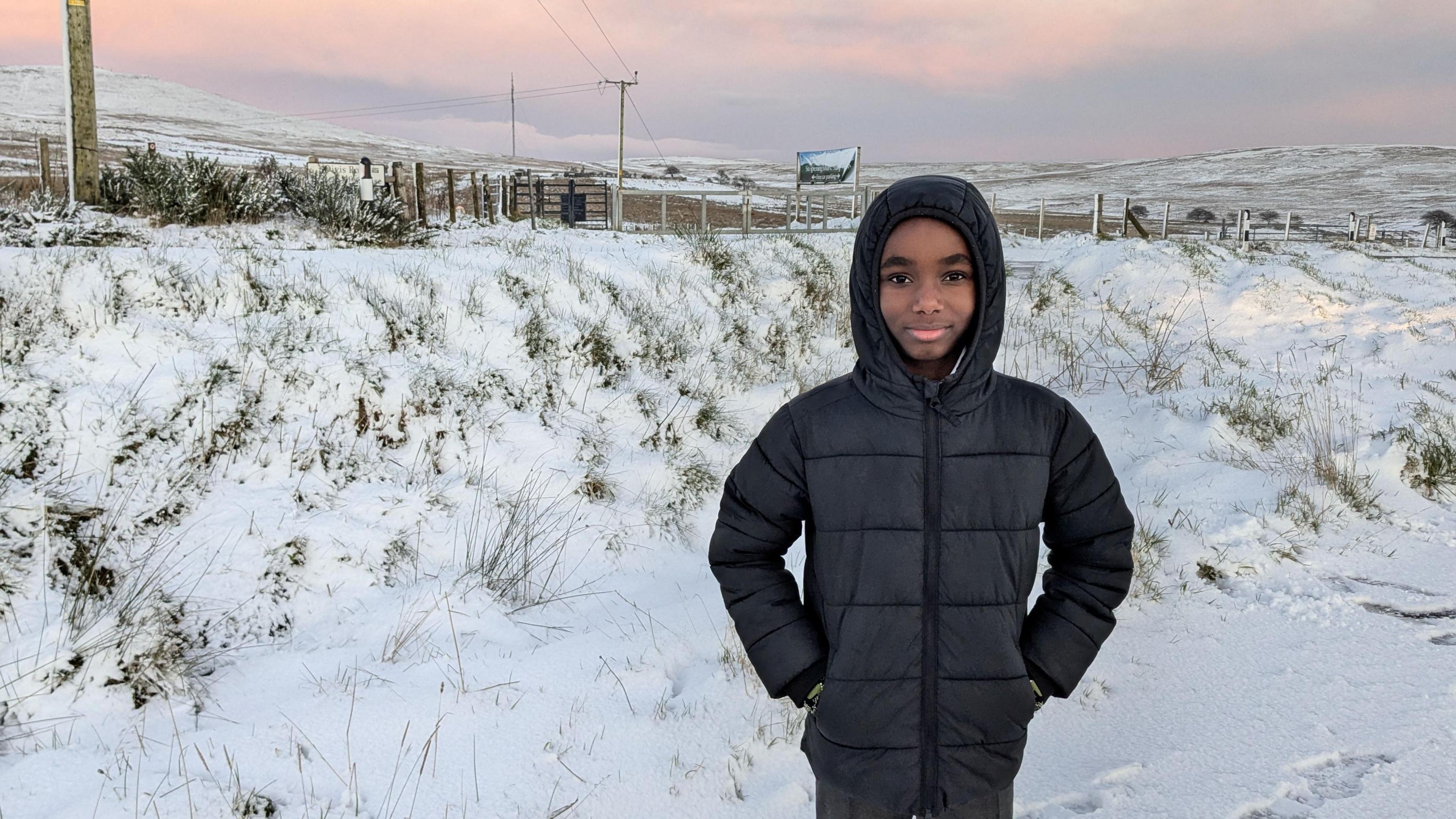 a boy in a thick puffer coat smiles into the camera, with his hands in his pockets. Around him the fields and hills are blanketed in a couple of centimetres of snow