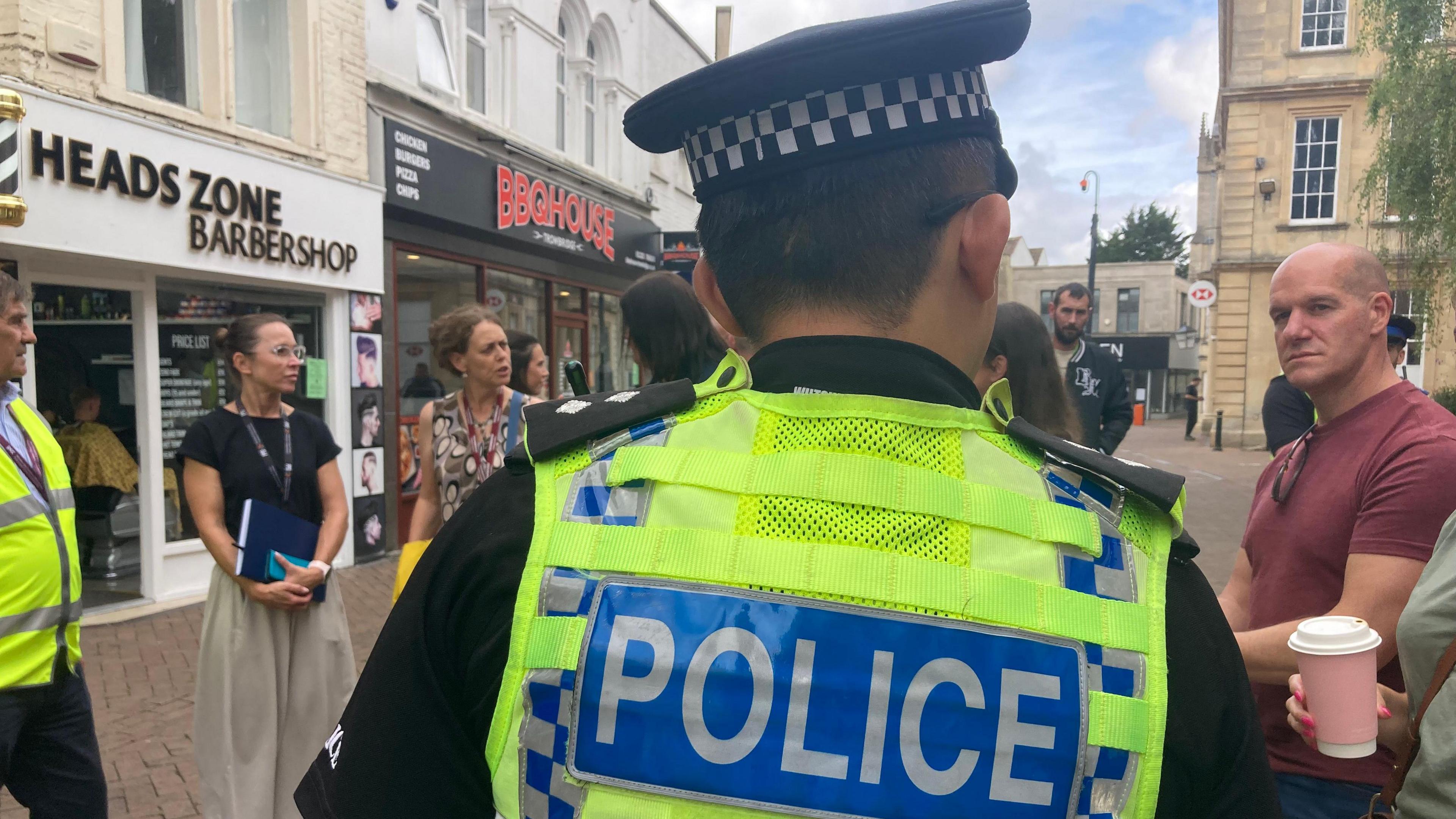 A police officer with his back to the camera. He is wearing a black police hat and a high vis vest with police written on the back. There are many people stood around him.
