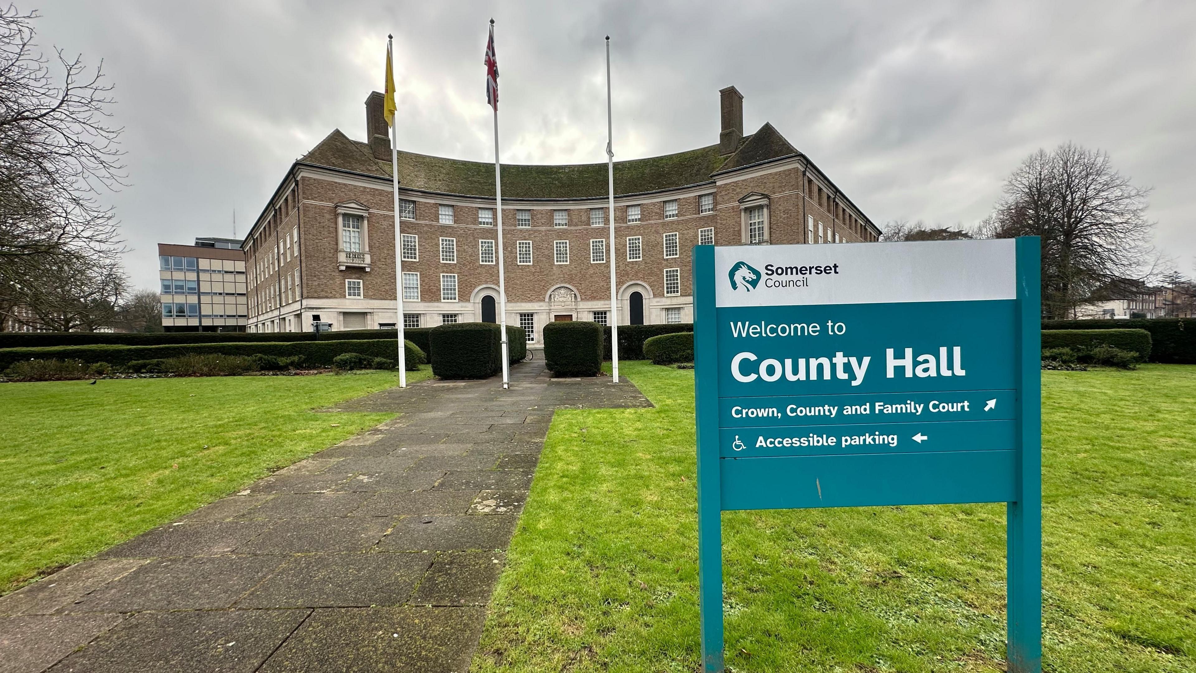 A turquoise sign reading welcome to county hall mounted in a lawn in front of a curved stone municipal building