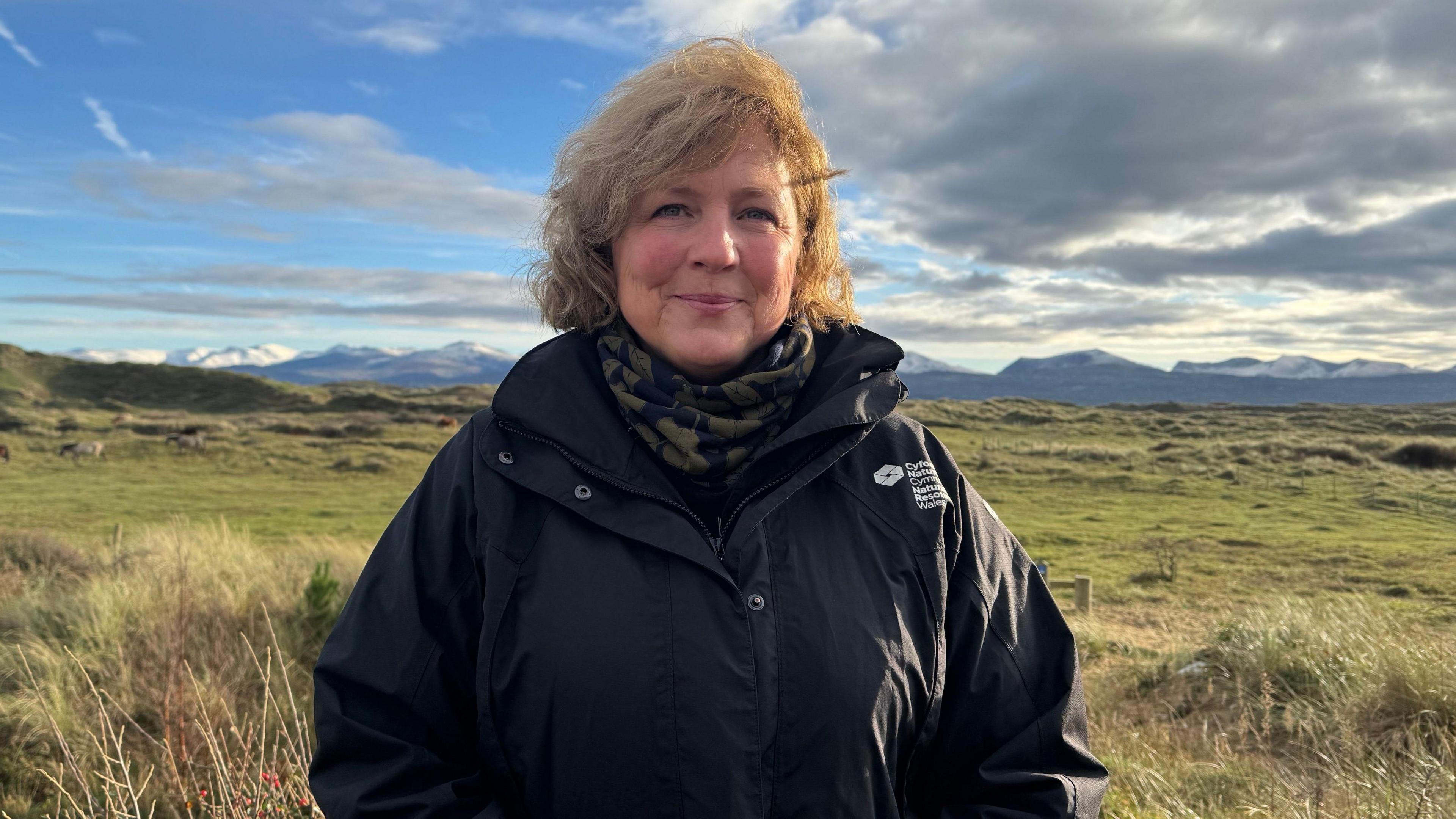 Portrait of Mannon Lewis, framed in the centre of the image. She has shoulder length wavey blonded or light brown hair and is smiling as she looks directly into the camera. She is wearing a black winter coat, with the white Natural Resources Wales logo in English and Welsh on the left side of the coast. She is wearing a scarf or neckwarmer, that is dark green and black. She is stood on pasture that is in fact sand dunes at Newborough Warren on Anglesey. Behind her can be seen the outline of mountains on the mainland, including the Carneddau and Eryri ranges, with some covered in snow. The sky is a mix of blue and some heavy dramatic clouds. It is a head and shoulders shot of her. 