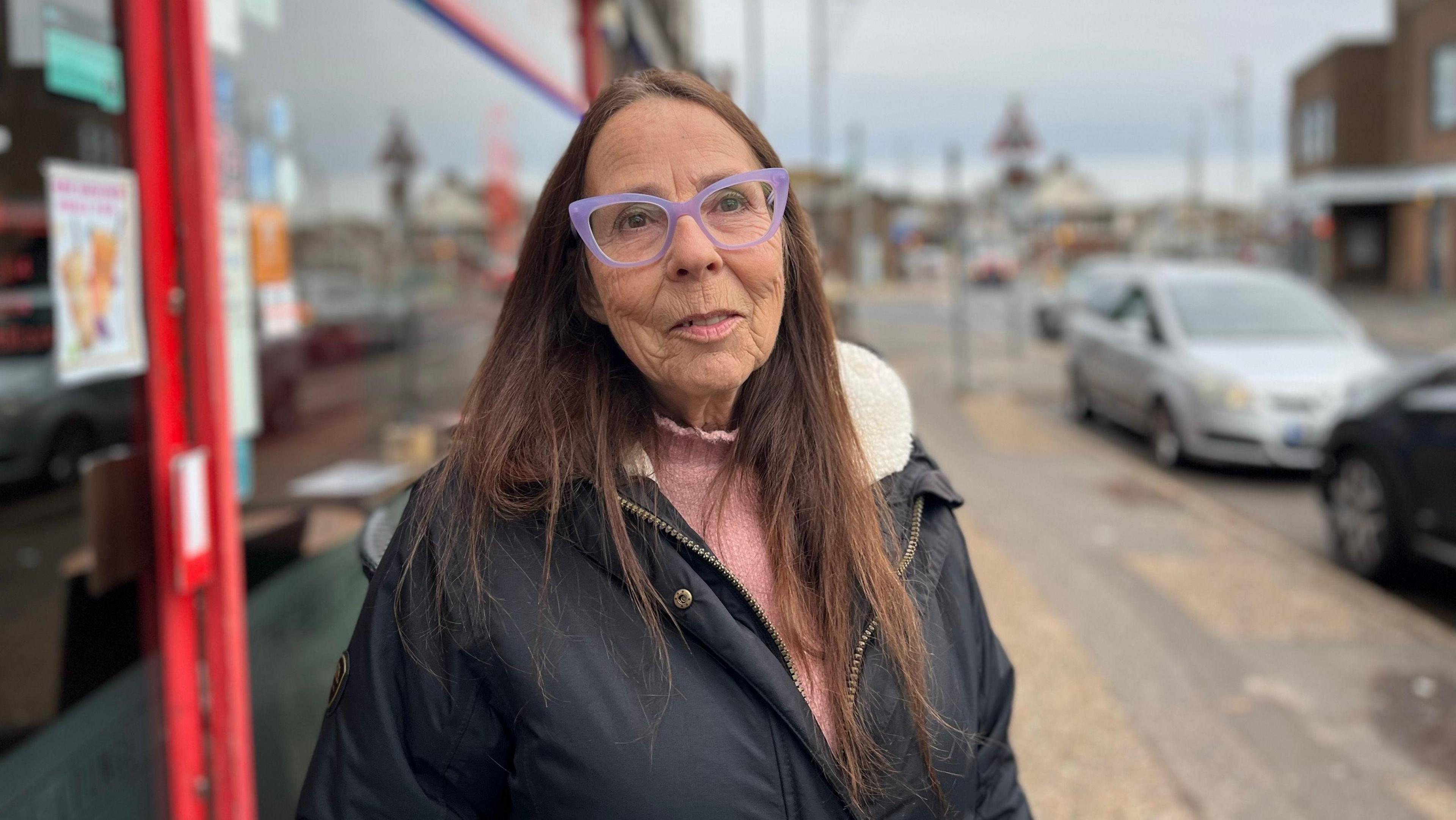 Joan Sloman who has long brown hair and purple glasses. She is wearing a winter coast and pink salmon jumper while standing on the pavement which has two cars parked next to her