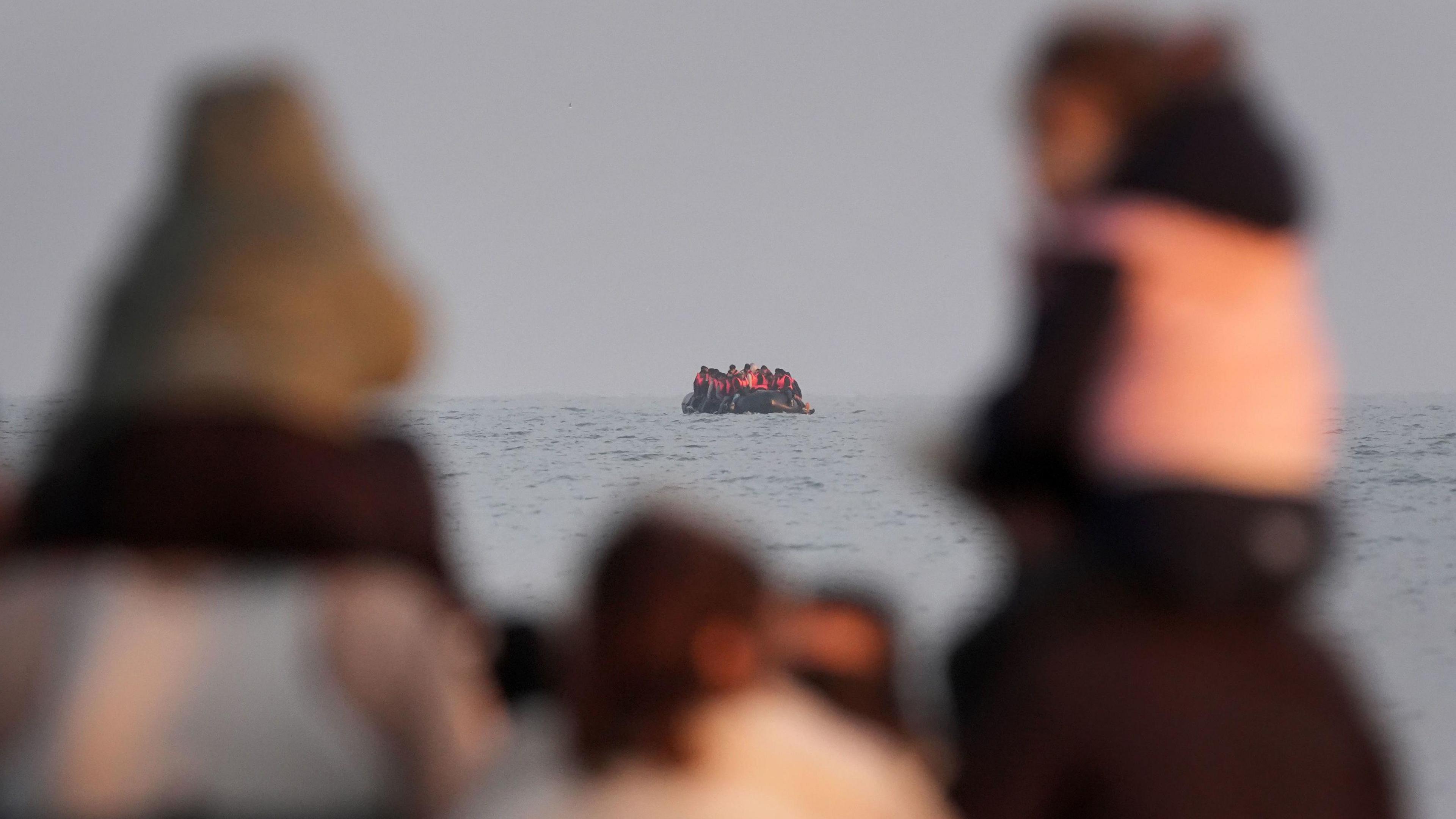 In the foreground are two children, carried on the shoulders of adults. They are pictured out of focus, looking out to sea. In the distance, a small boat full of people wearing life jackets is seen at sea.